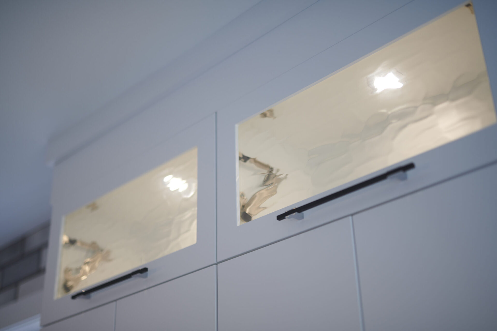 An image showcasing a modern white kitchen cabinet with glossy, cream-colored doors featuring black handles. The ceiling light reflects off the doors' surface.