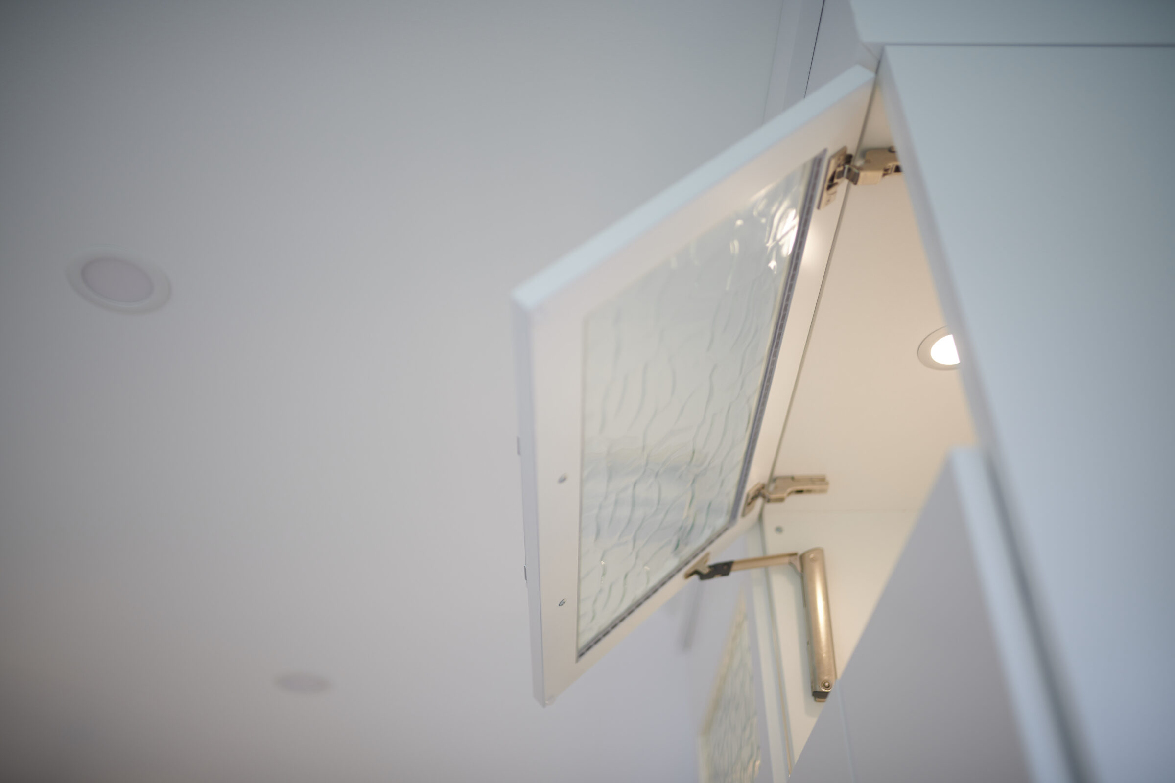 An open textured glass window set in a white wall, hinged at the top, with a bright ceiling light visible in the soft-focus background.