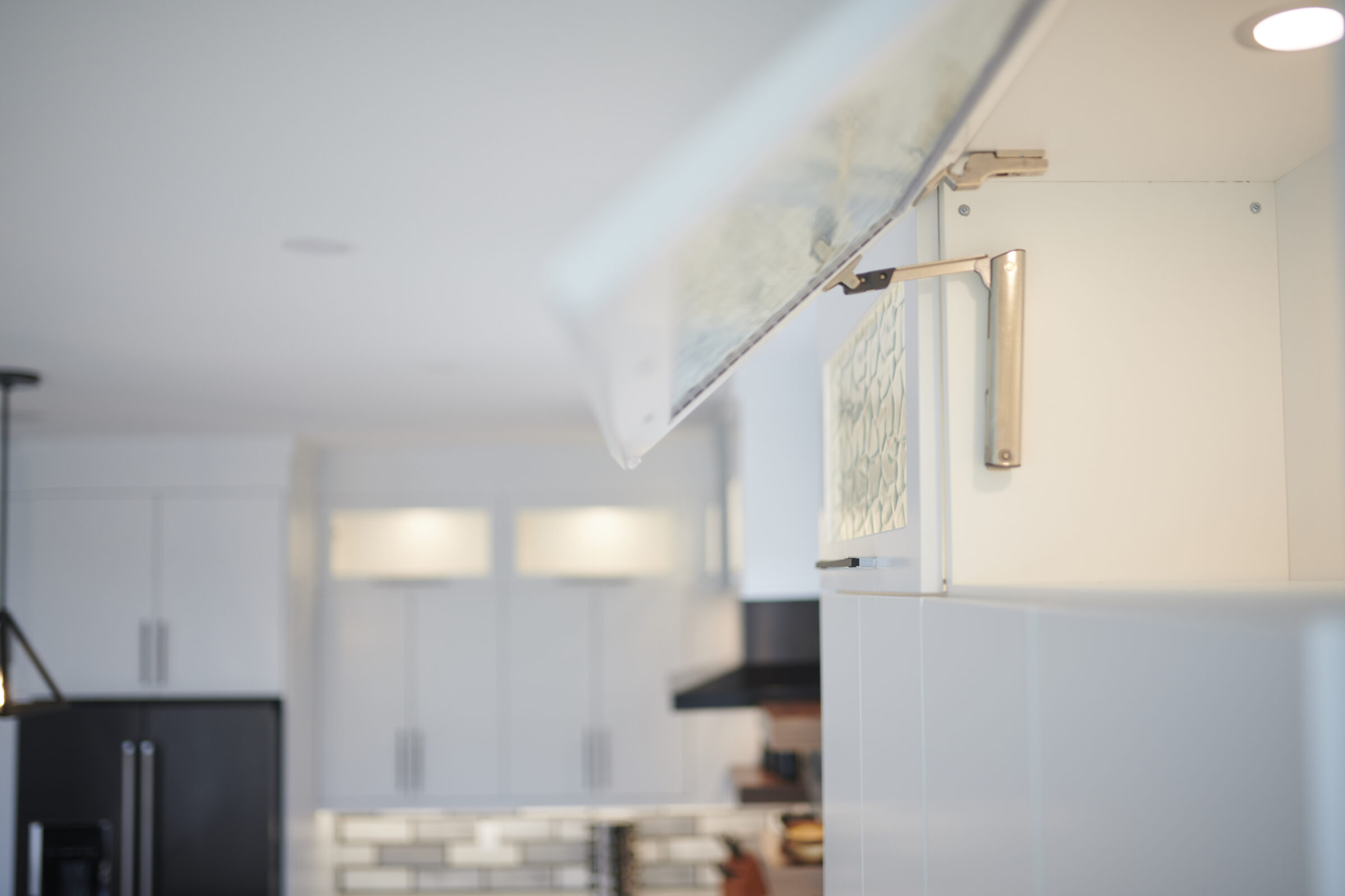 This is a softly lit, modern kitchen with white cabinetry. An overhead cabinet door is open, showing hinges; blurred background reveals more of the interior.