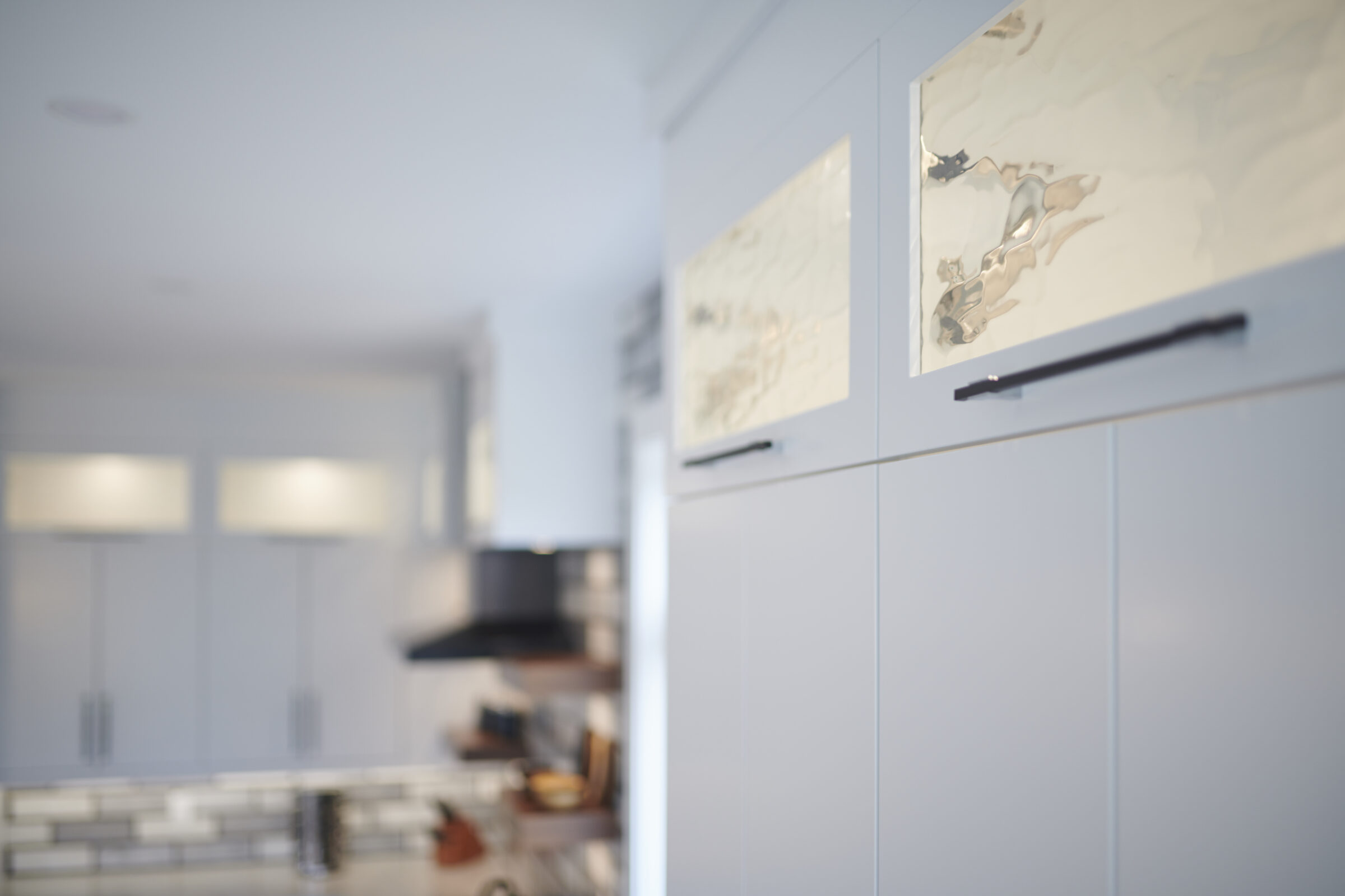 This image shows a blurred modern kitchen with white cabinetry and handles, focused on metallic decorative elements on the cabinet doors.