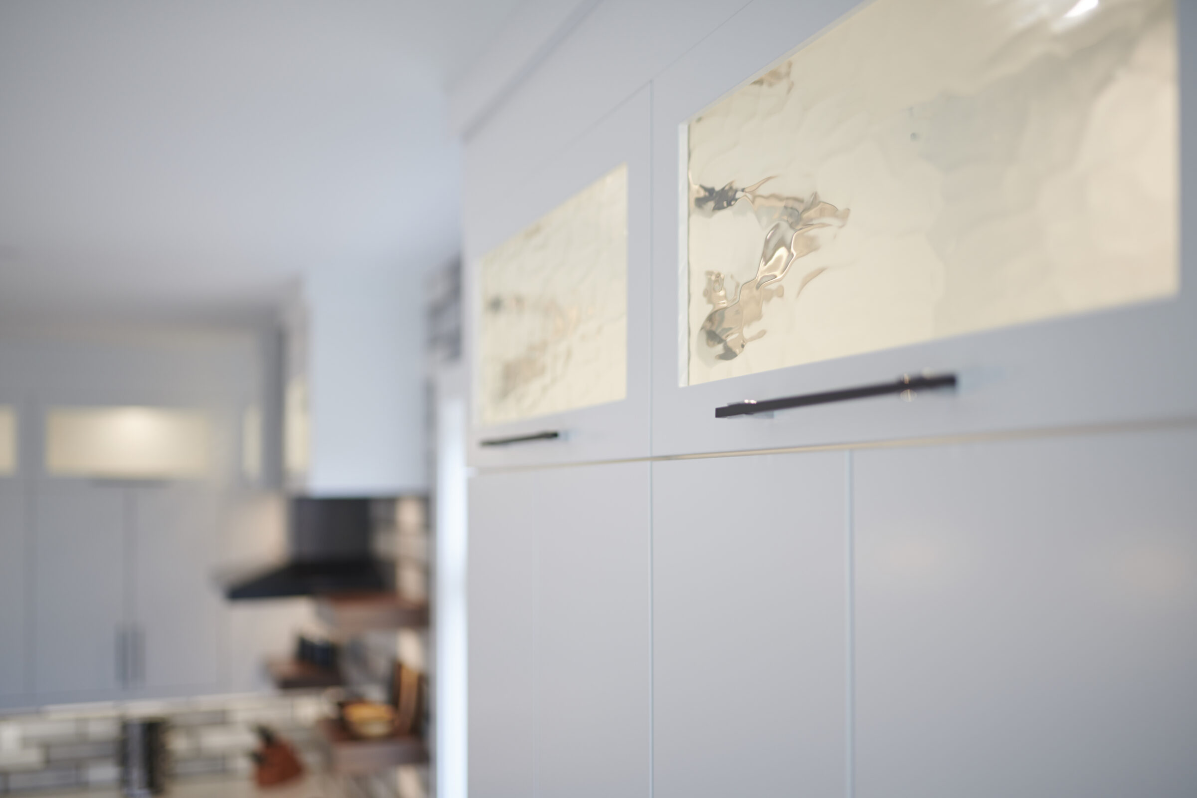 This is a modern kitchen with white cabinets. Artwork hangs above, and the room has a blurred background with some wooden elements visible.