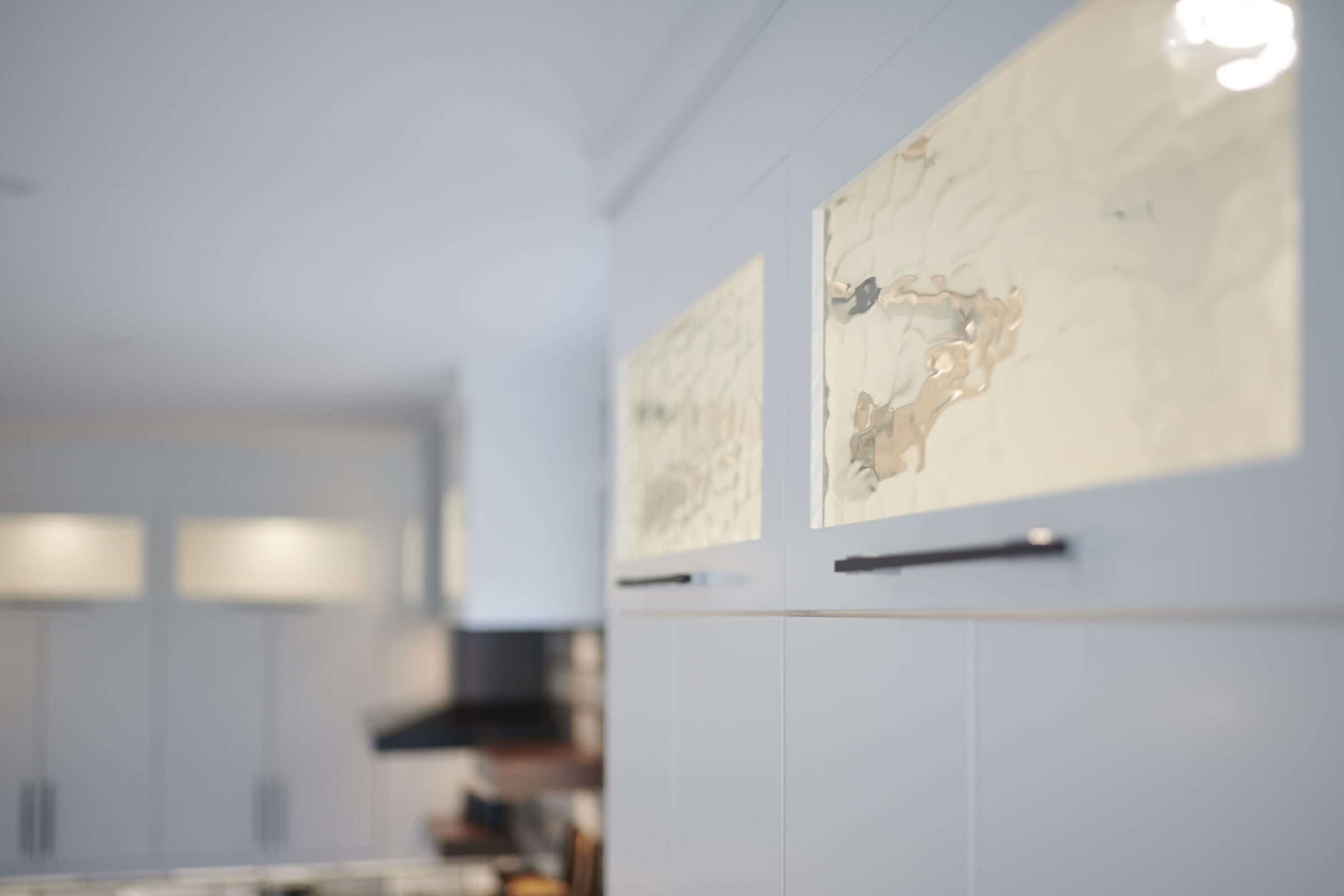 Modern kitchen interior with blurred focus. White cabinets with metallic handles, reflective backsplashes. Clean, minimalist design with soft lighting. No people visible.
