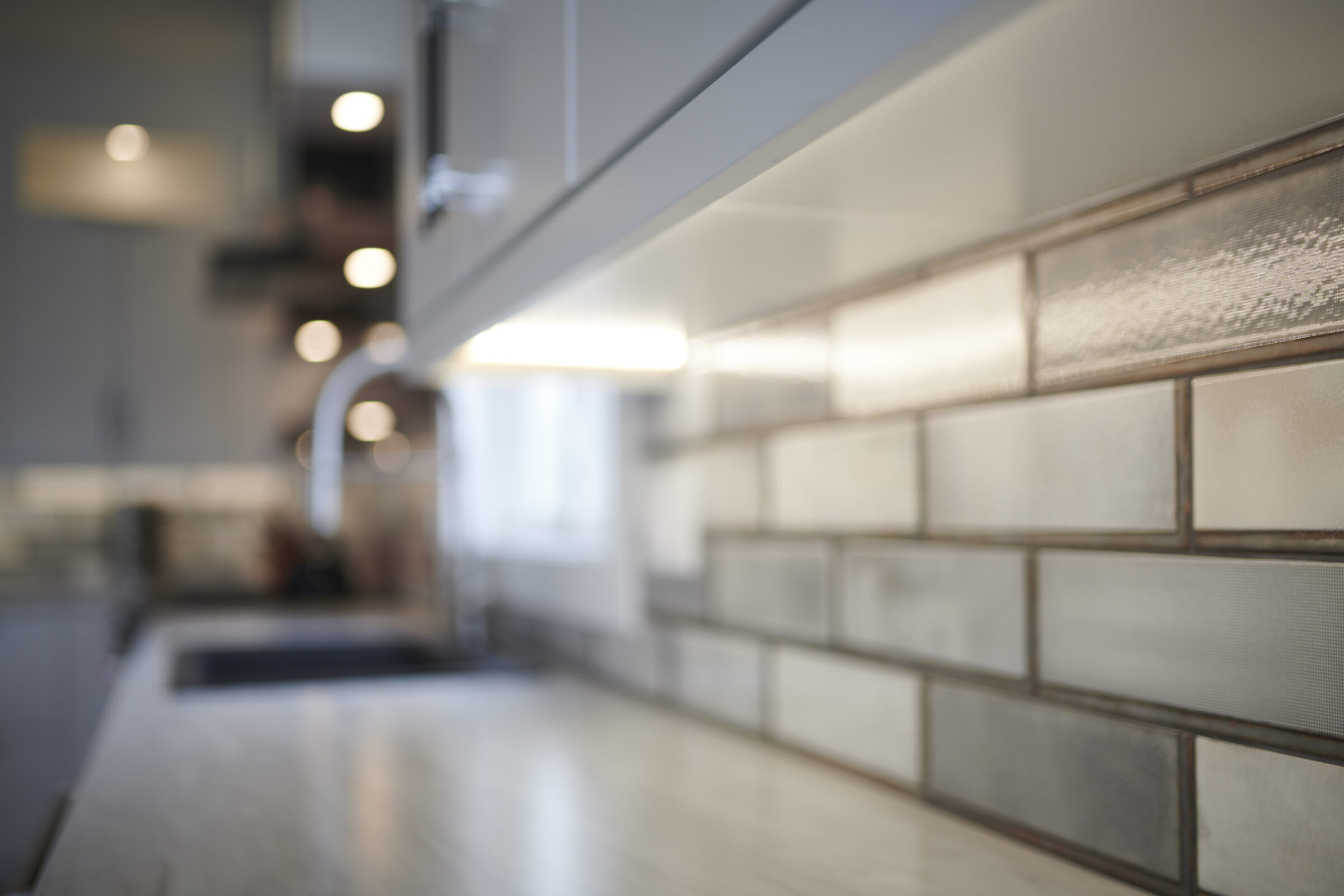 A blurred kitchen interior focuses on textured subway tiles with a defocused background showing light fixtures, countertops, and a faint silhouette of a person.