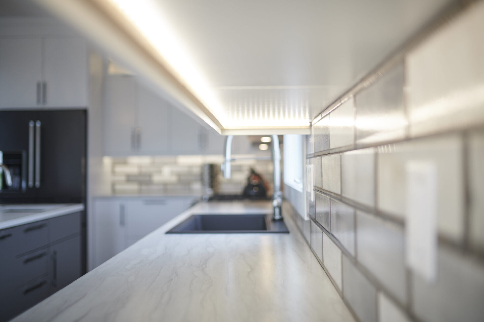 This is a modern kitchen with a focus on a marble countertop, under-cabinet lighting, and subway tile backsplash, with blurred background appliances.