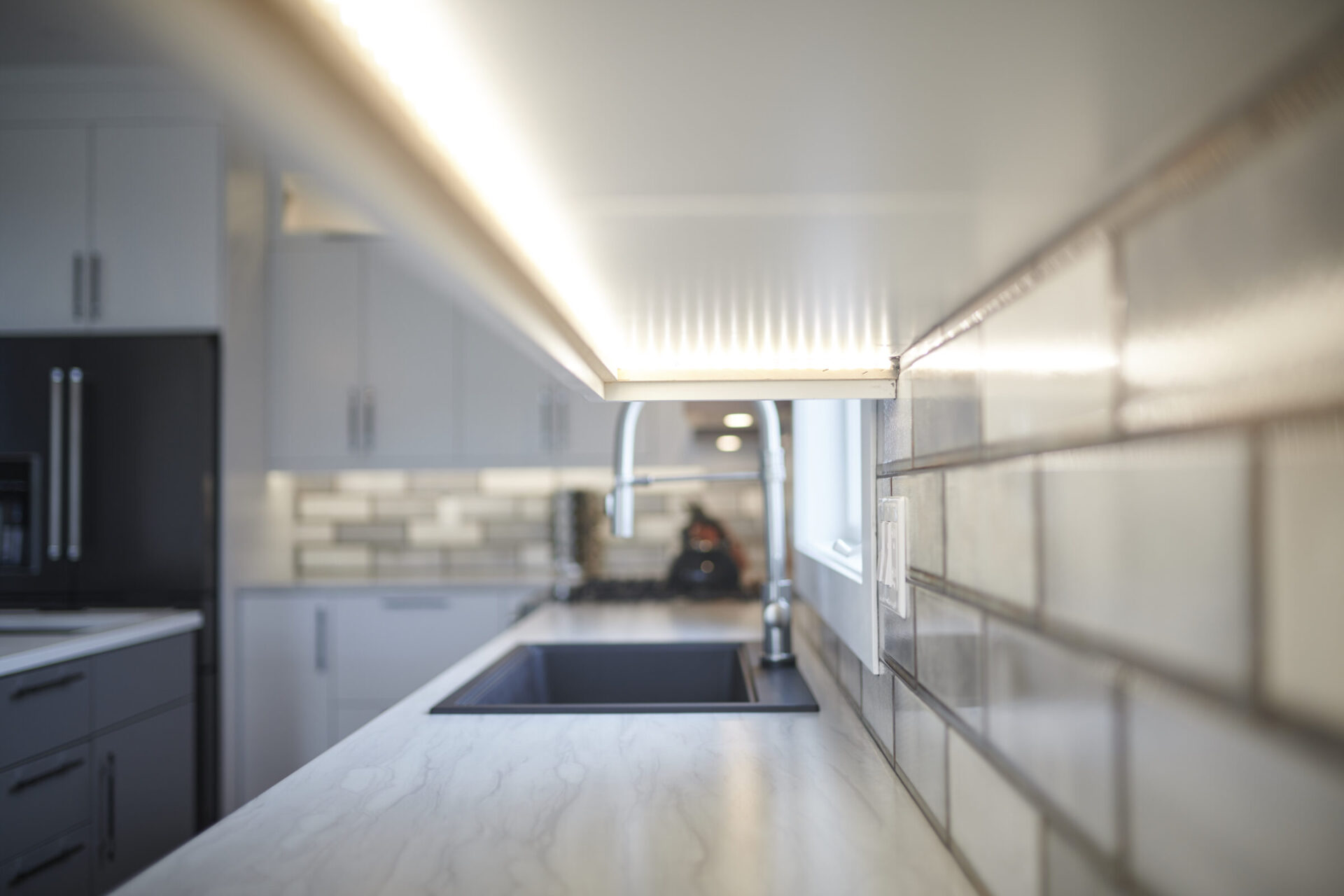 Modern kitchen interior with under-cabinet lighting, tiled backsplash, marble countertop, and a blurred background hinting at sleek appliances. Focus is on the light reflection.