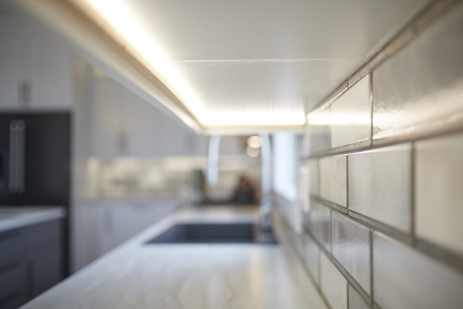 This image features an out-of-focus modern kitchen with sleek surfaces, a strip of bright lighting above, and subway tiles on the wall.