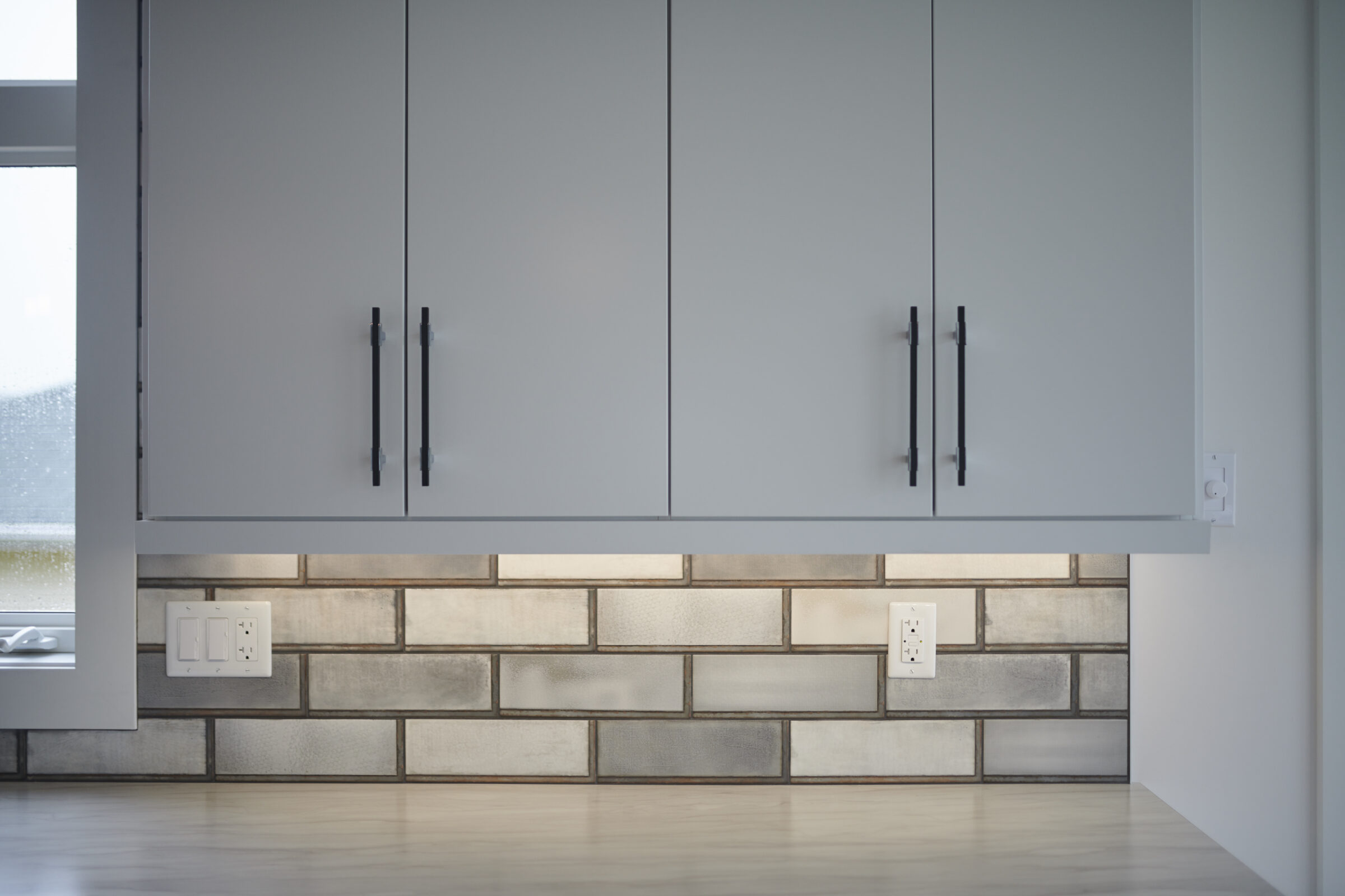 A modern kitchen with grey cabinets, metallic handles, a subway tile backsplash, wood flooring, and white electrical outlets on the wall.