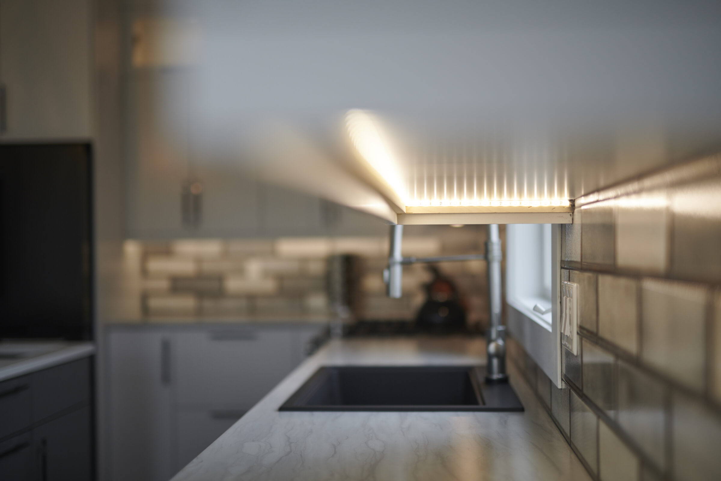 Modern kitchen interior focused on a countertop under an illuminated cabinet, with blurry grey cabinetry and backsplash tiles in the background.