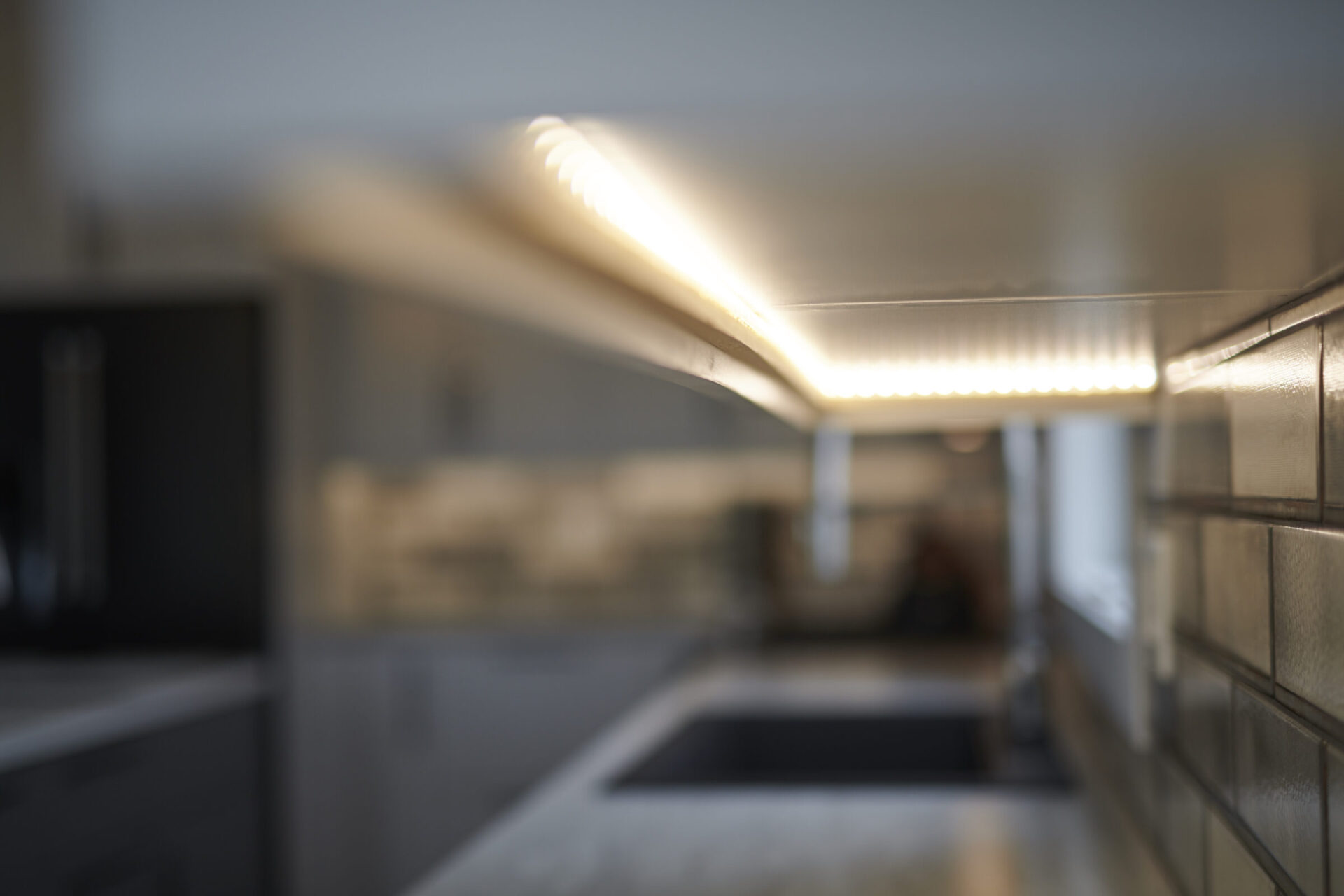 This image shows an out-of-focus interior, possibly a kitchen, with a clear focus on a lit, elongated LED light fixture against a tiled wall.