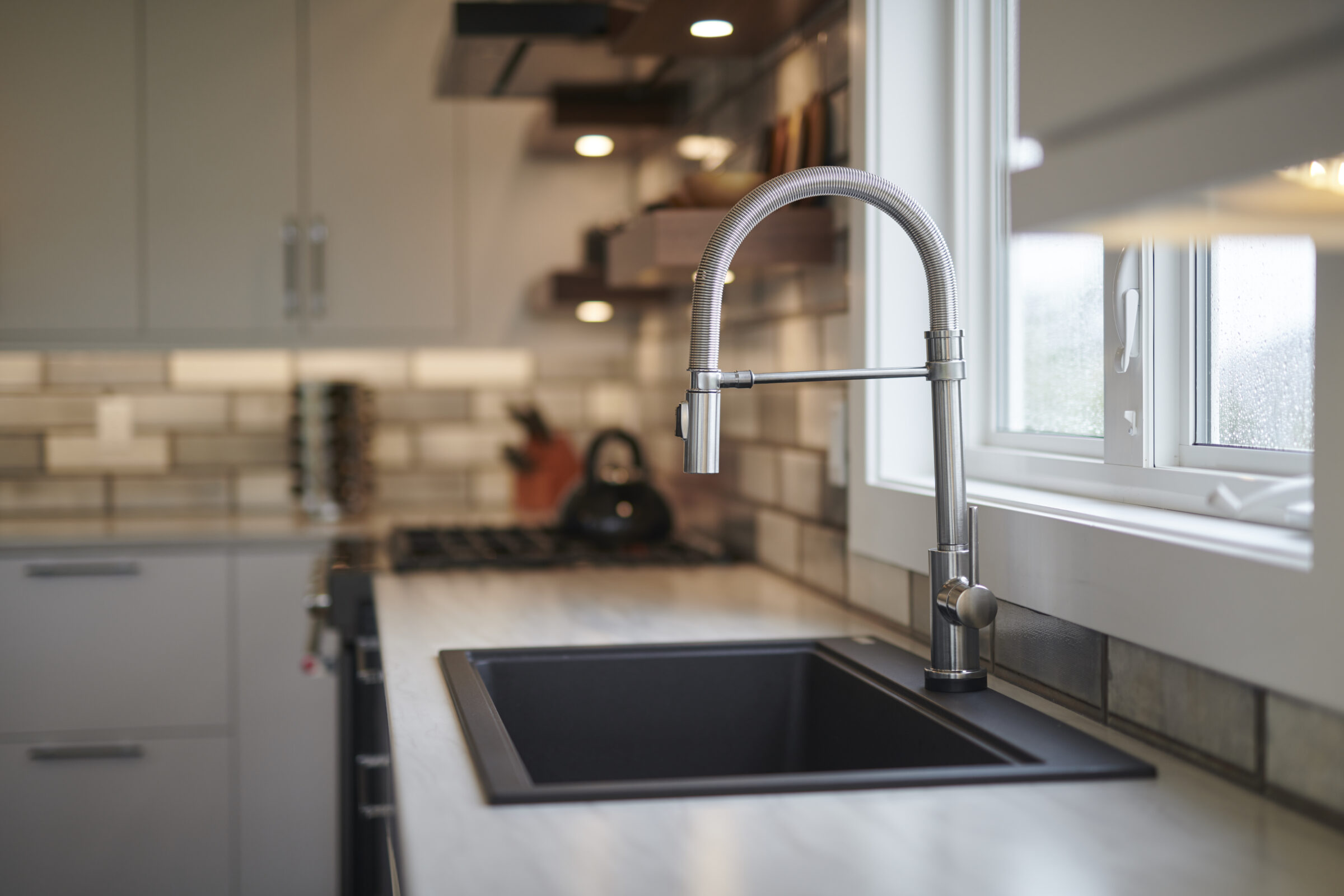 Modern kitchen interior featuring a stainless steel sink and flexible faucet, with light beige tile backsplash, under a window with raindrops visible outside.