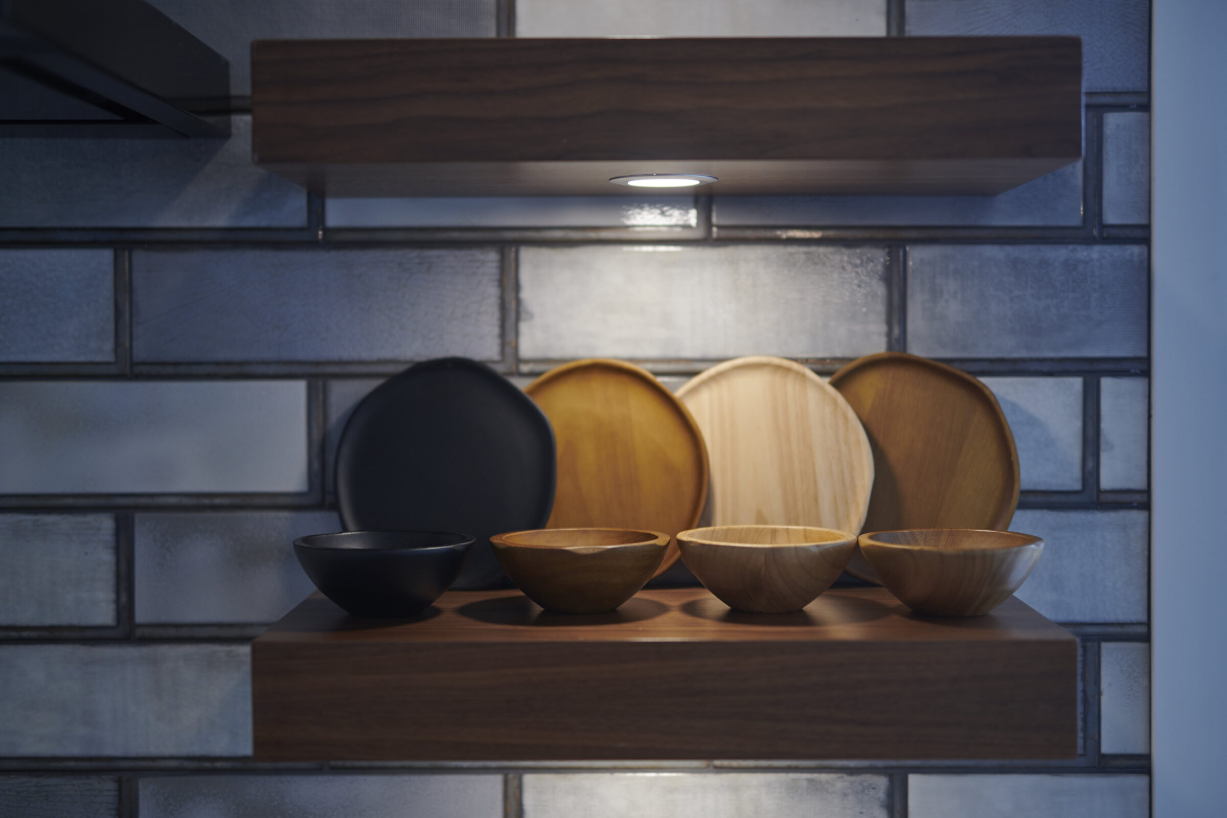 A modern kitchen shelf with assorted wooden and black bowls and plates. The shelf is underlit and set against a tiled wall.