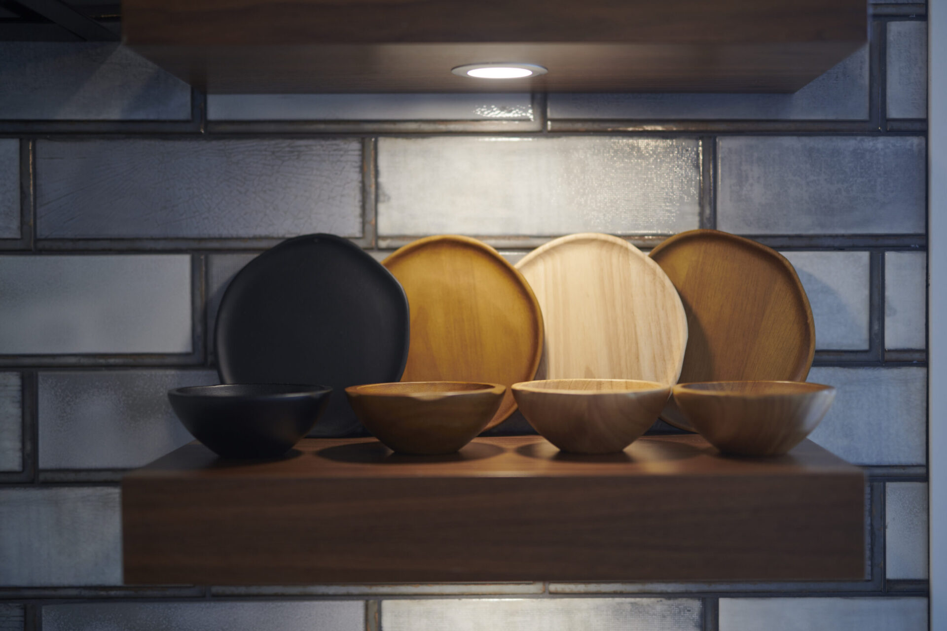 A wooden shelf holds various plates and bowls against a blue-tiled backdrop with a warm, illuminated ambiance from a small shelf light.