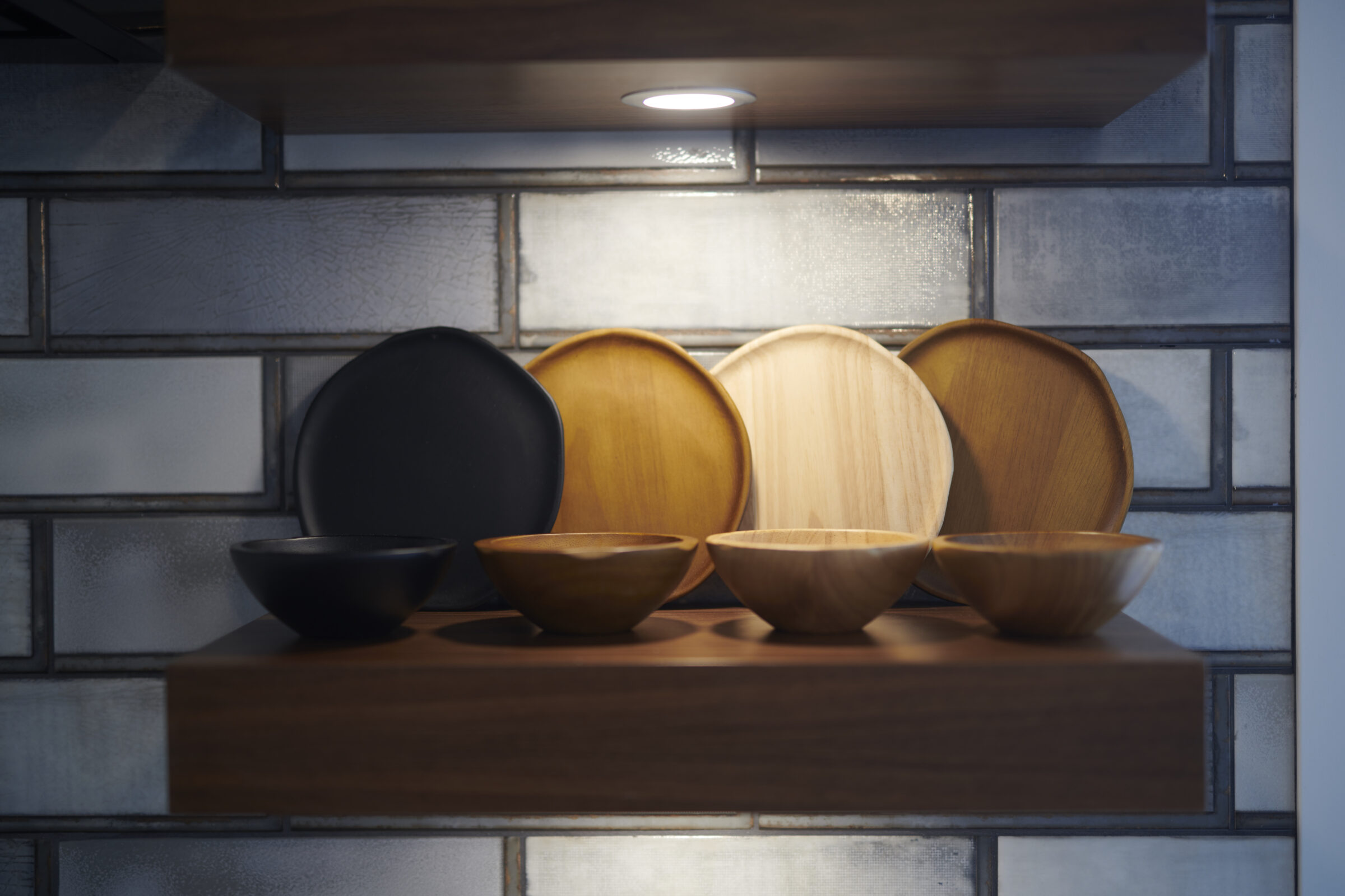 A selection of black and wooden plates and bowls arranged on a shelf with a spotlight above, against a blue-grey tiled backsplash.