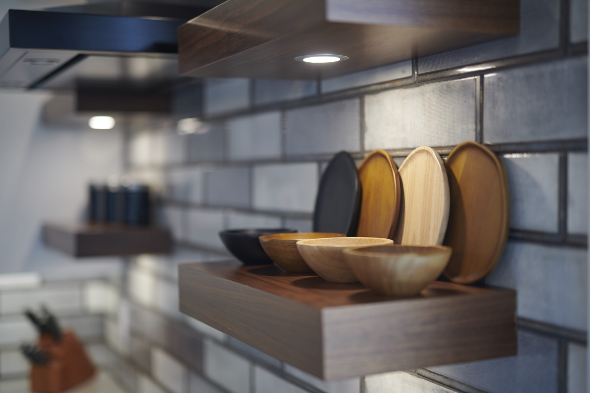 A modern kitchen shelf holds a variety of wooden bowls against a backsplash of grey tiles, with blurred kitchenware in the background.