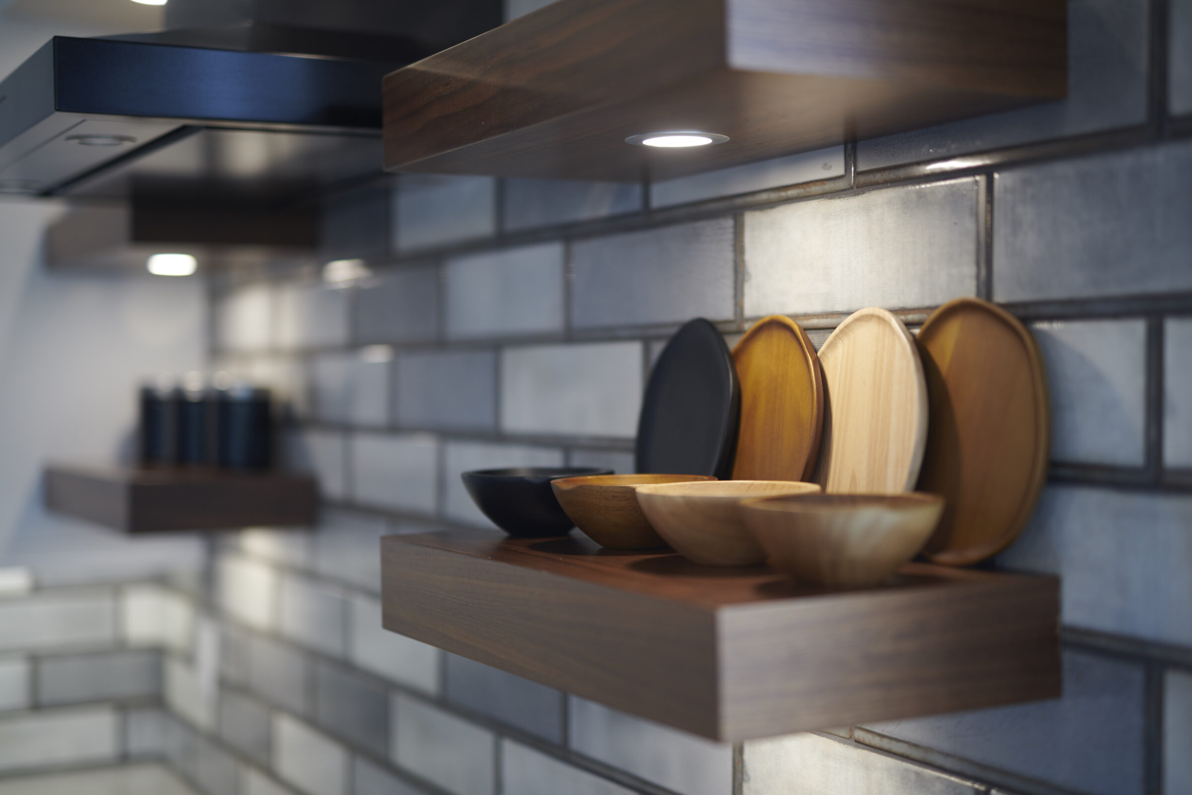 A modern kitchen setting featuring wooden shelves with a neat arrangement of variously colored bowls against a gray tiled backsplash under cabinet lighting.
