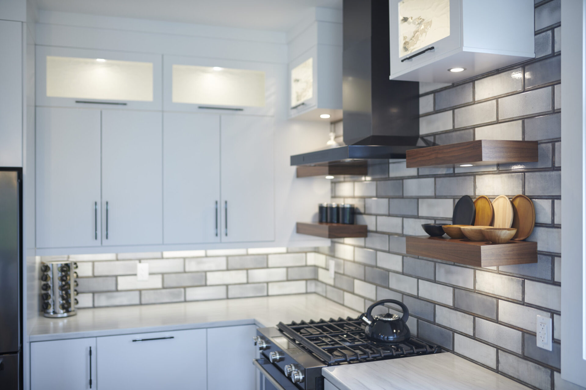 A modern kitchen interior with white cabinetry, floating wooden shelves, a gas stove, subway tiles backsplash, and a teapot on the countertop.