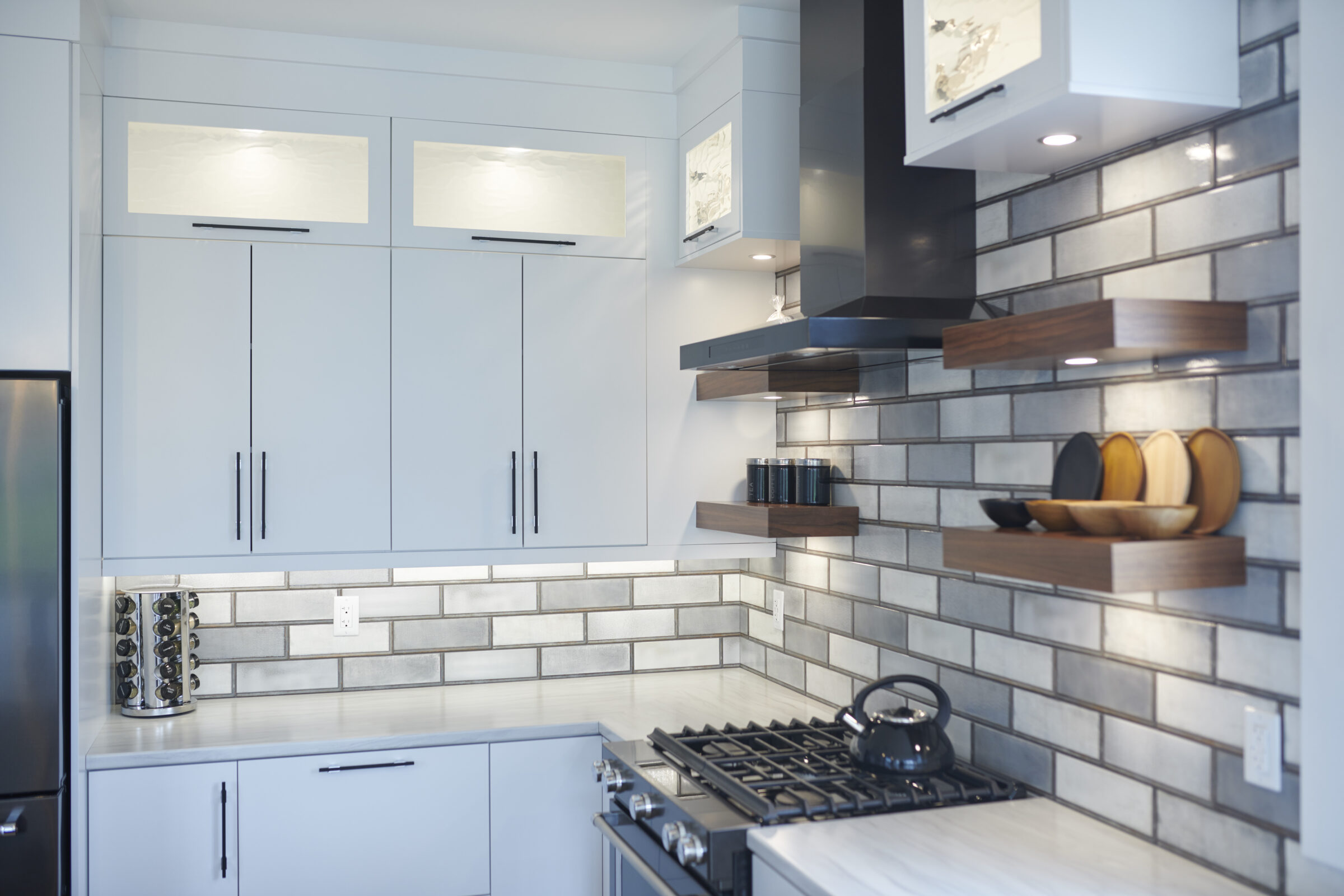 Modern kitchen corner with stainless steel gas stove, white cabinets, floating wooden shelves, a black kettle, and grey subway tile backsplash.