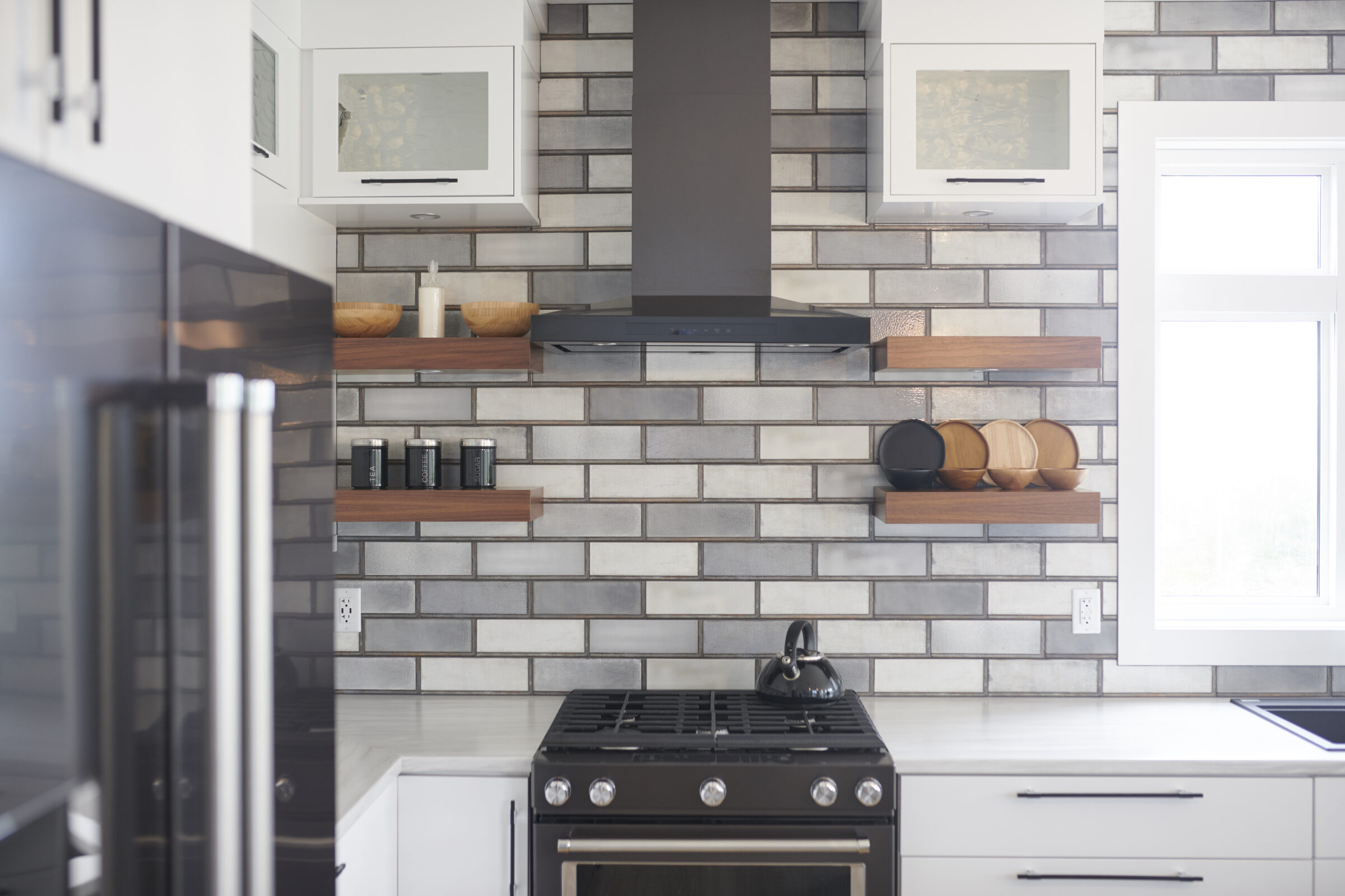 A modern kitchen with stainless steel appliances, white cabinets, gray subway tiles, floating wooden shelves, and cookware on display near a window.