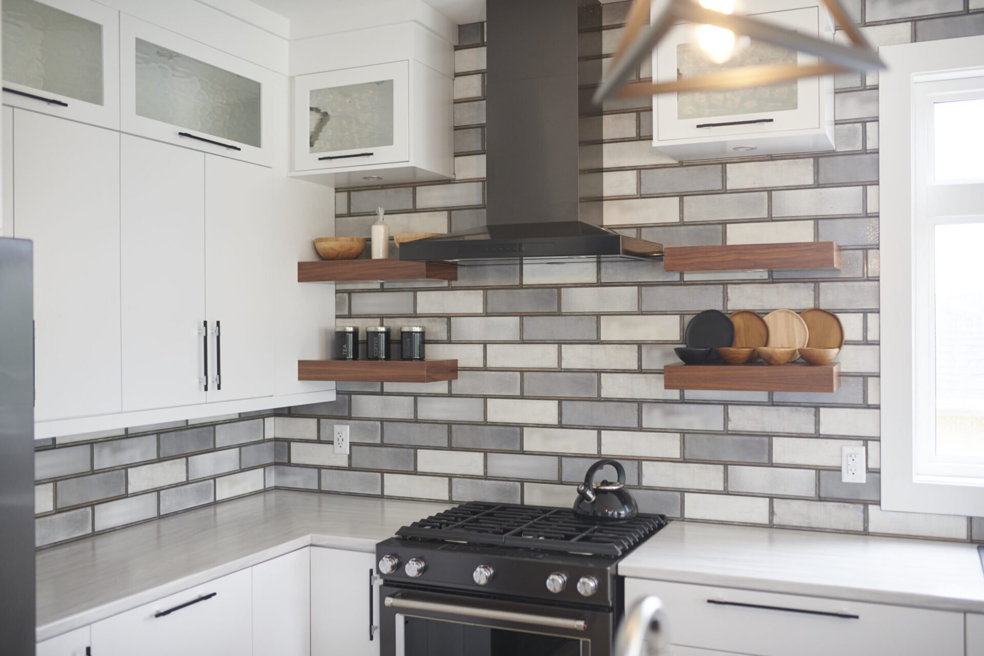 A modern kitchen with white cabinets, stainless steel appliances, floating wooden shelves, brick-style backsplash, and a kettle on the stove.