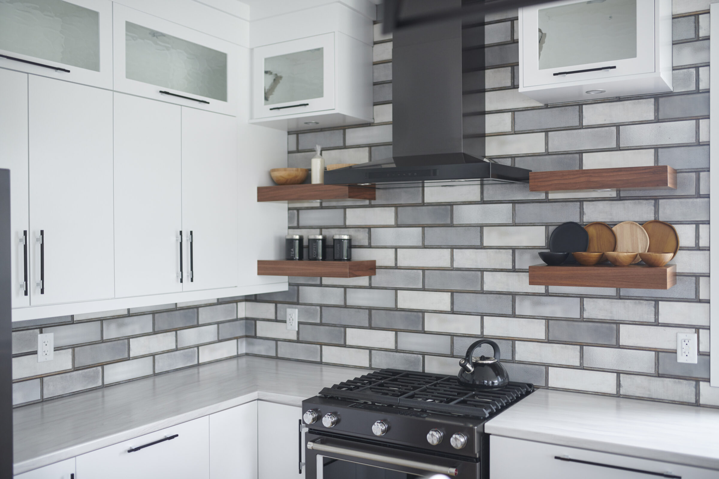 A modern kitchen with stainless steel appliances, white cabinets, grey subway tile backsplash, and wooden floating shelves holding utensils and containers.