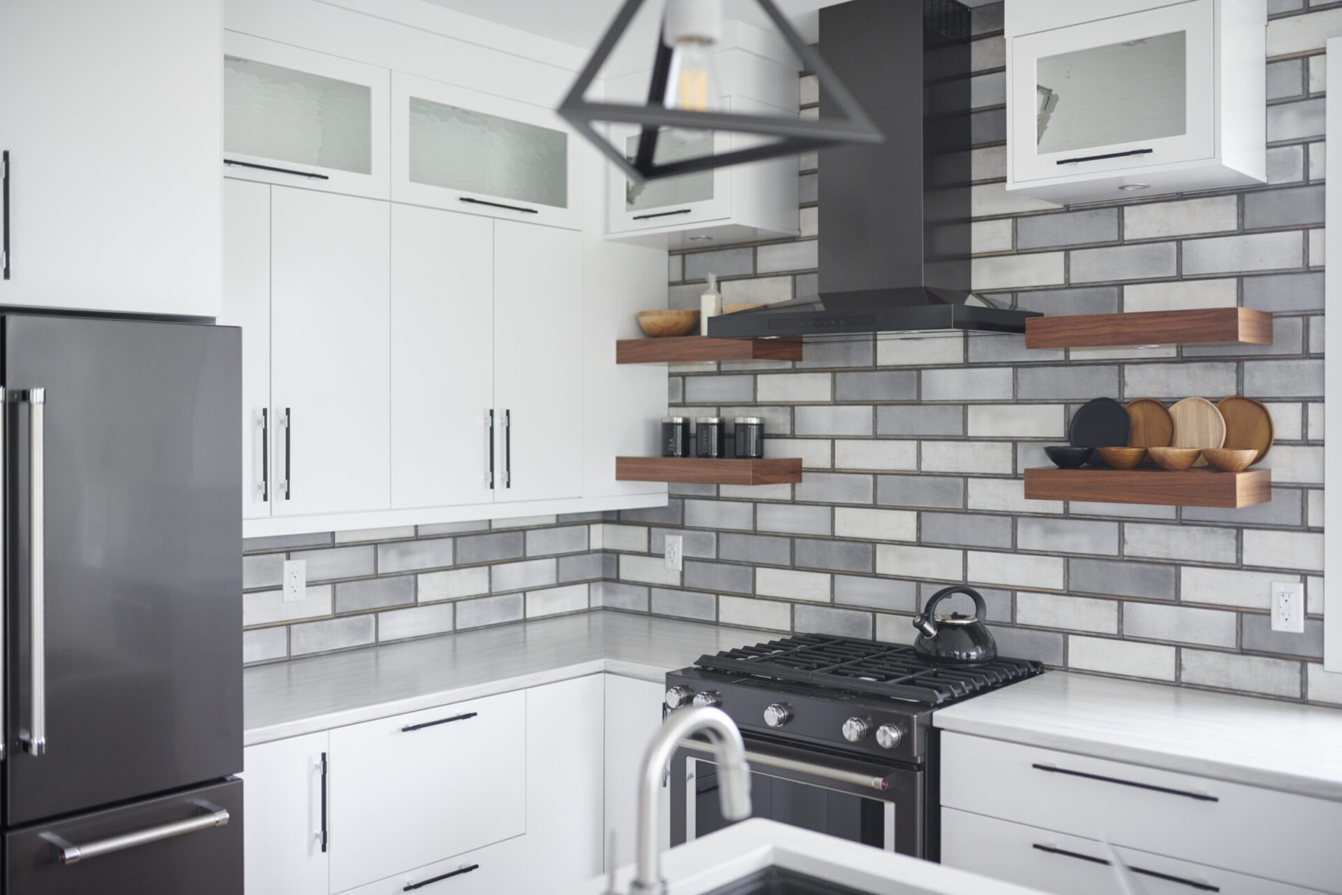 Modern kitchen interior with stainless steel appliances, white cabinets, gray subway tile backsplash, wooden shelves, and a geometric light fixture.