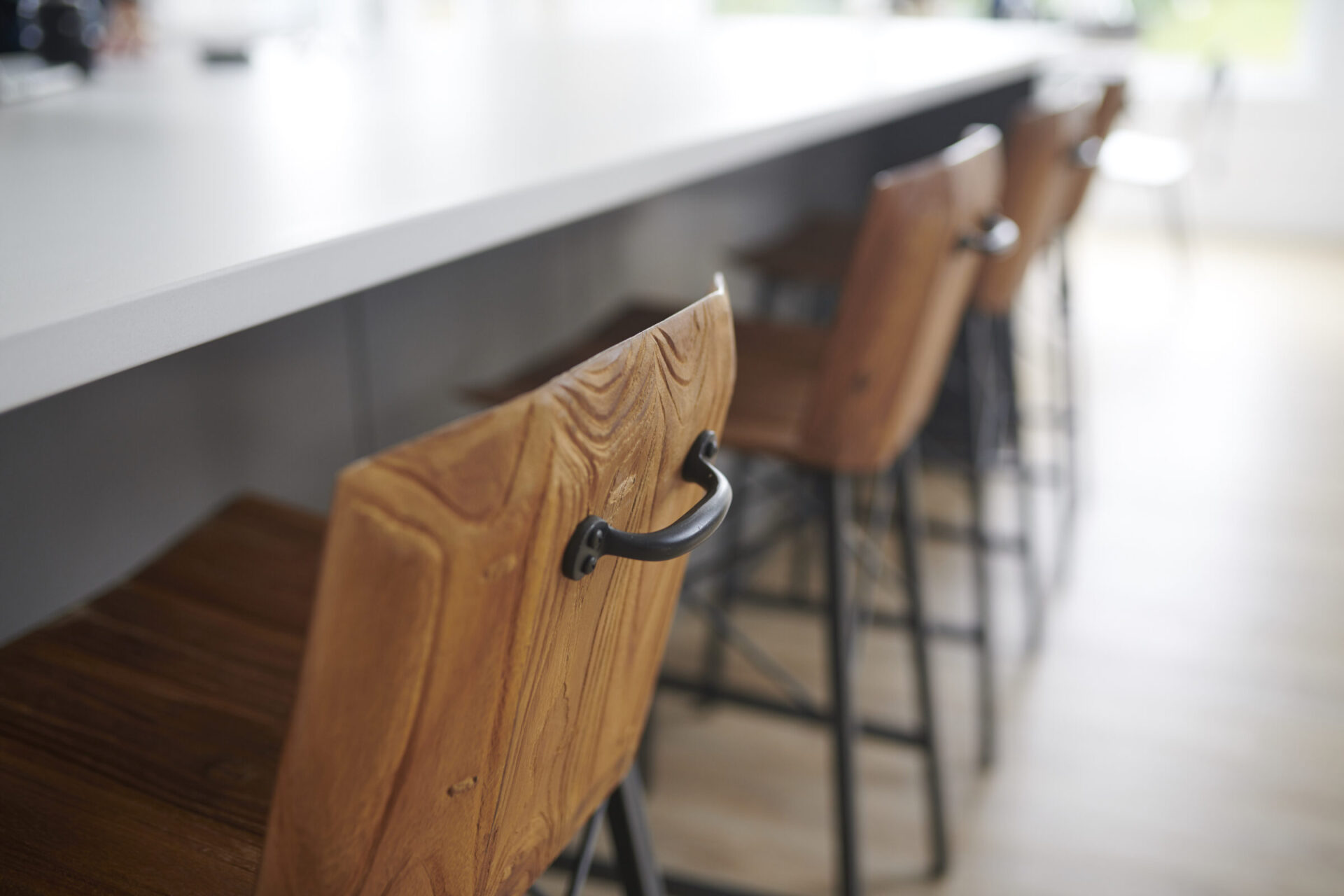 A modern kitchen bar with stylish wooden stools, black metal legs, set against a white countertop and a blurred background with bright lighting.