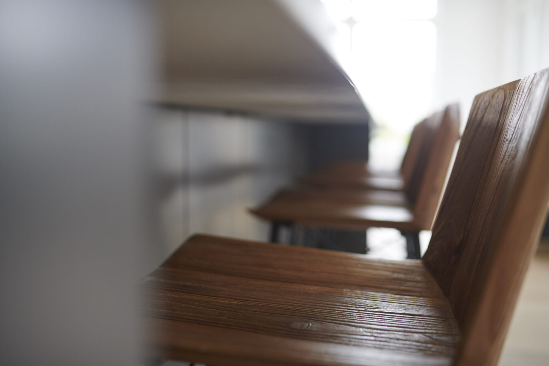 A row of wooden chairs against a white wall with focused lighting; a shallow depth of field blurs the background, highlighting texture and form.