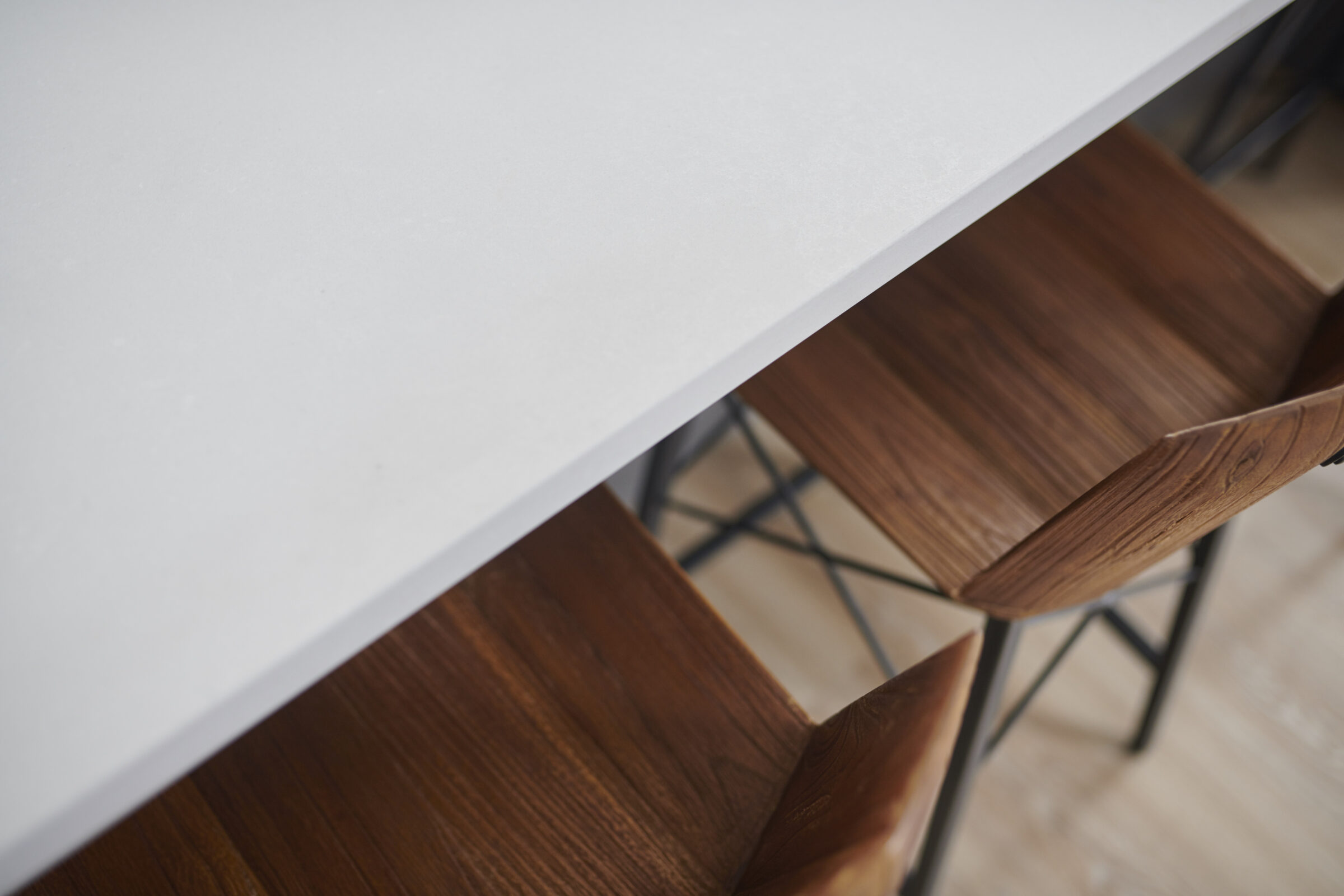 A minimalist setting featuring a white tabletop with sleek edges above wooden chairs with metal legs, arranged neatly on a hardwood floor.
