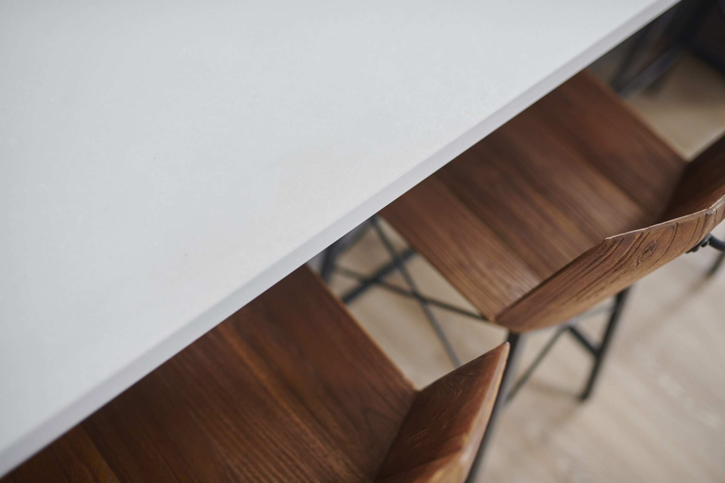 The image shows a close-up of a white tabletop with two brown wooden chairs below, featuring a minimalist and contemporary design aesthetic.