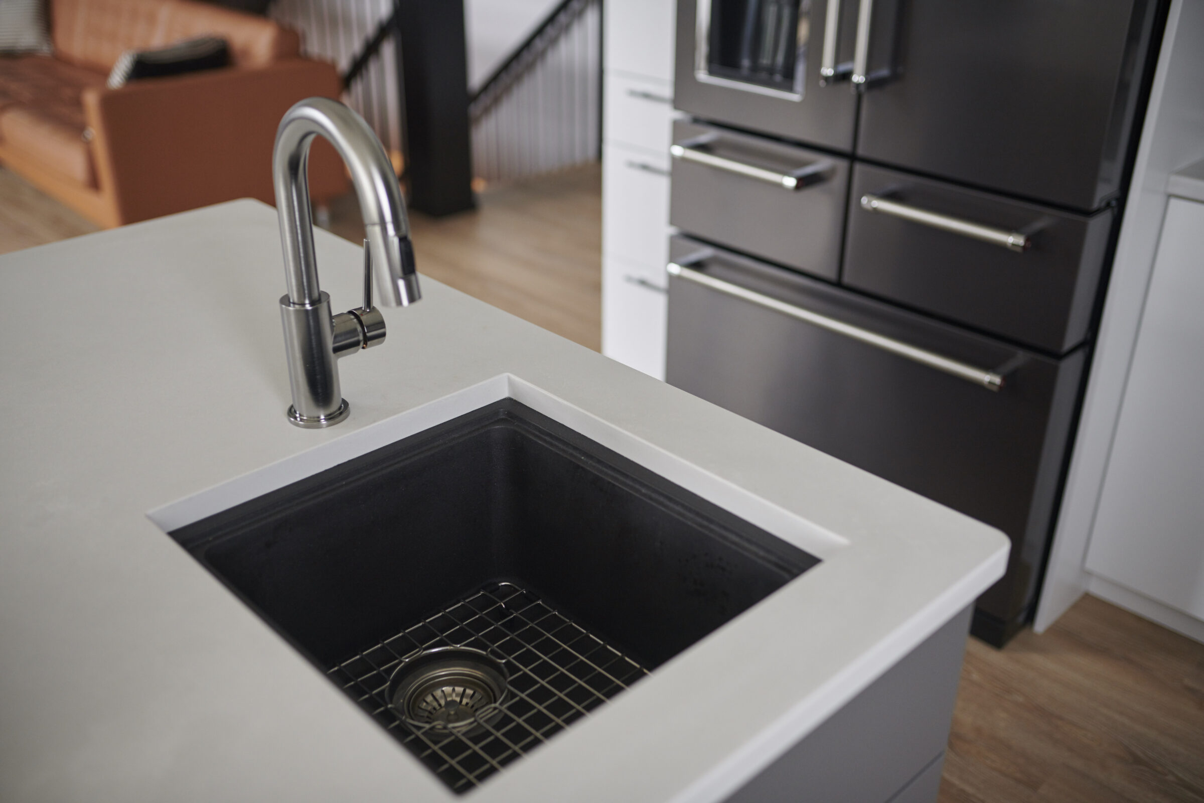 A modern kitchen featuring a black sink with a stainless steel faucet, white countertop, dark cabinetry, and a glimpse of a living area.