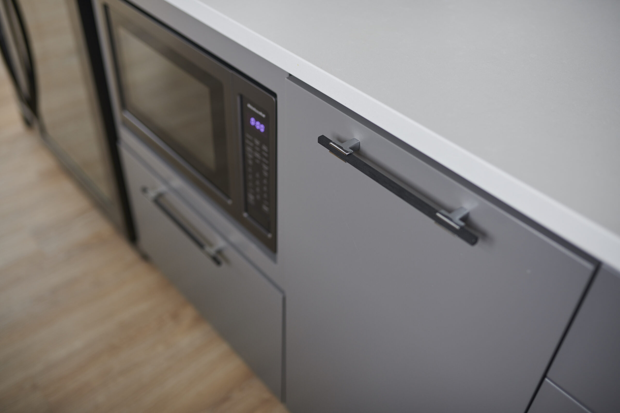 A modern kitchen featuring a built-in microwave and oven with sleek black handles set against gray cabinets and a light wood floor.