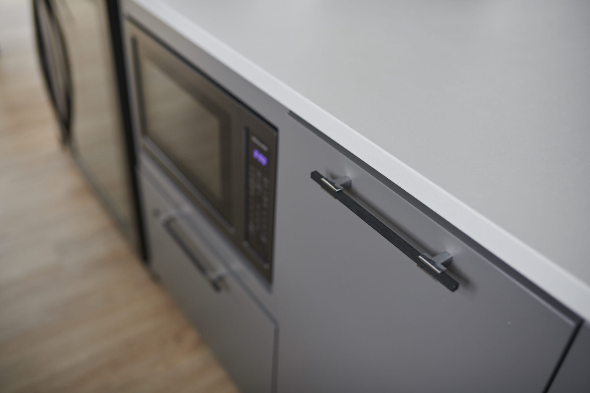 Modern kitchen with built-in stainless steel microwave and oven, sleek gray cabinetry with black handles, and wooden flooring. Focus is slightly blurred.