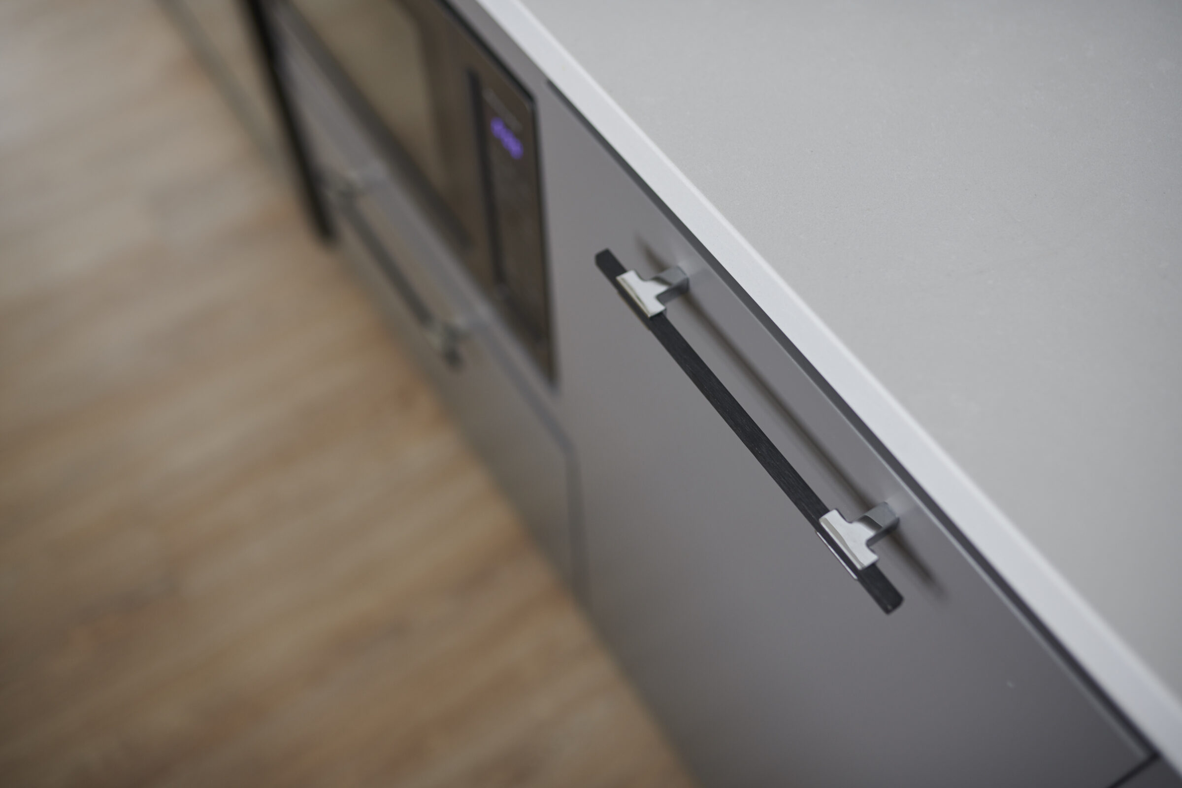 Modern kitchen with minimalist design. Gray cabinets have sleek black handles. Focused foreground with softly blurred background of wood flooring.