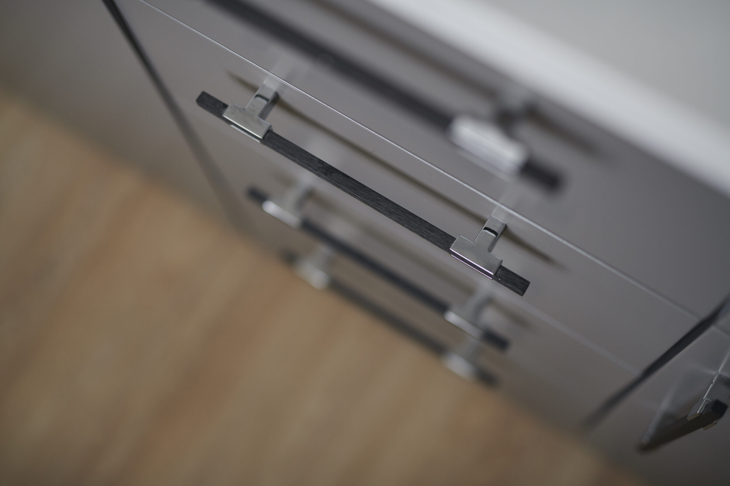 This image shows a modern, gray cabinet with sleek metal handles against a blurred wooden floor background, likely in a contemporary home interior.