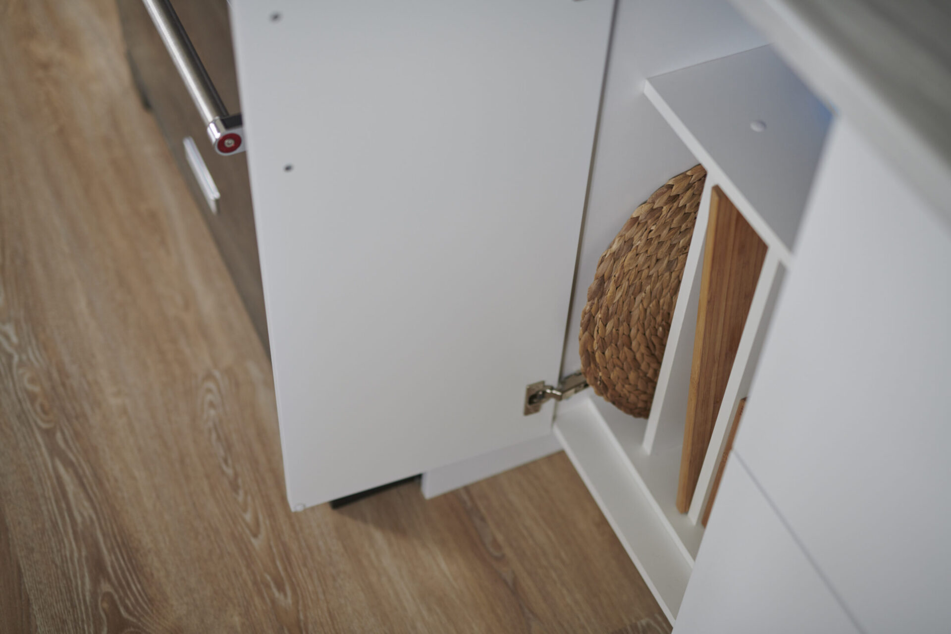 A partially open white kitchen cupboard with a metal handle, revealing a woven basket inside, against a wood laminate flooring background.