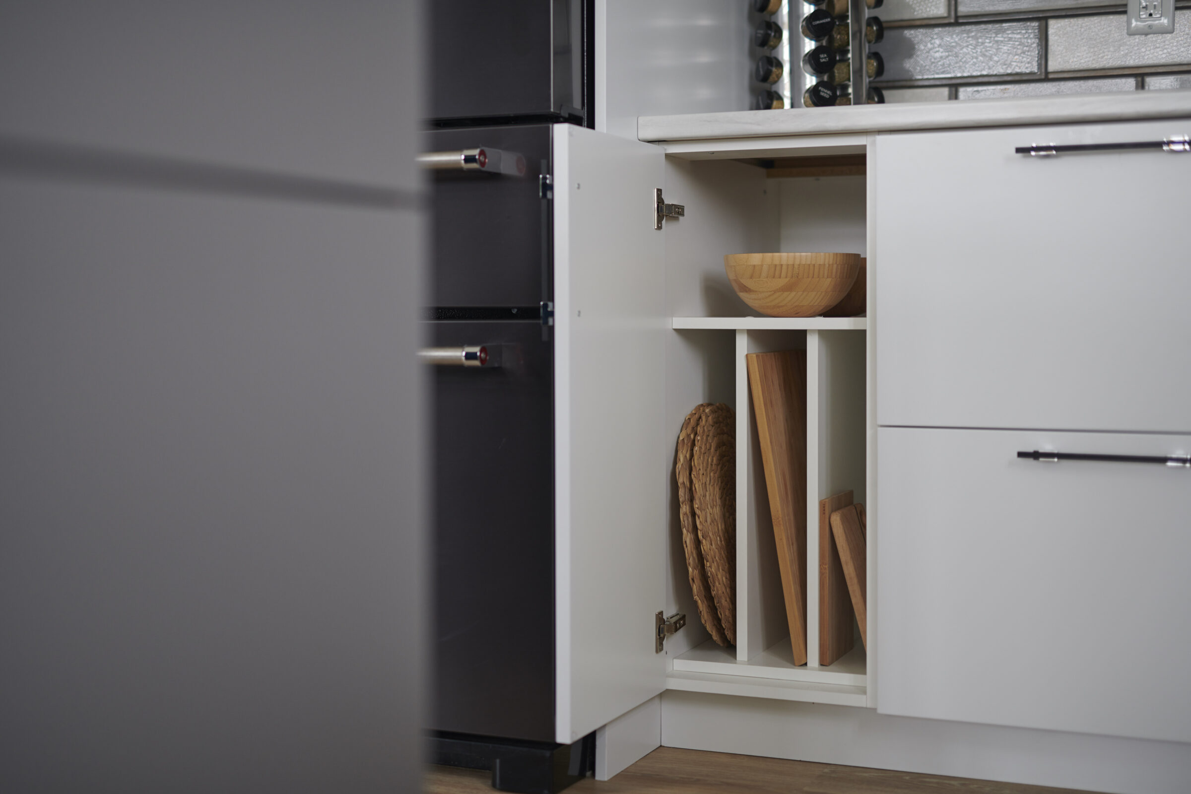 An open kitchen cabinet with shelves displaying a wooden bowl and cutting boards. A wine rack and refrigerator are partially visible to the left.