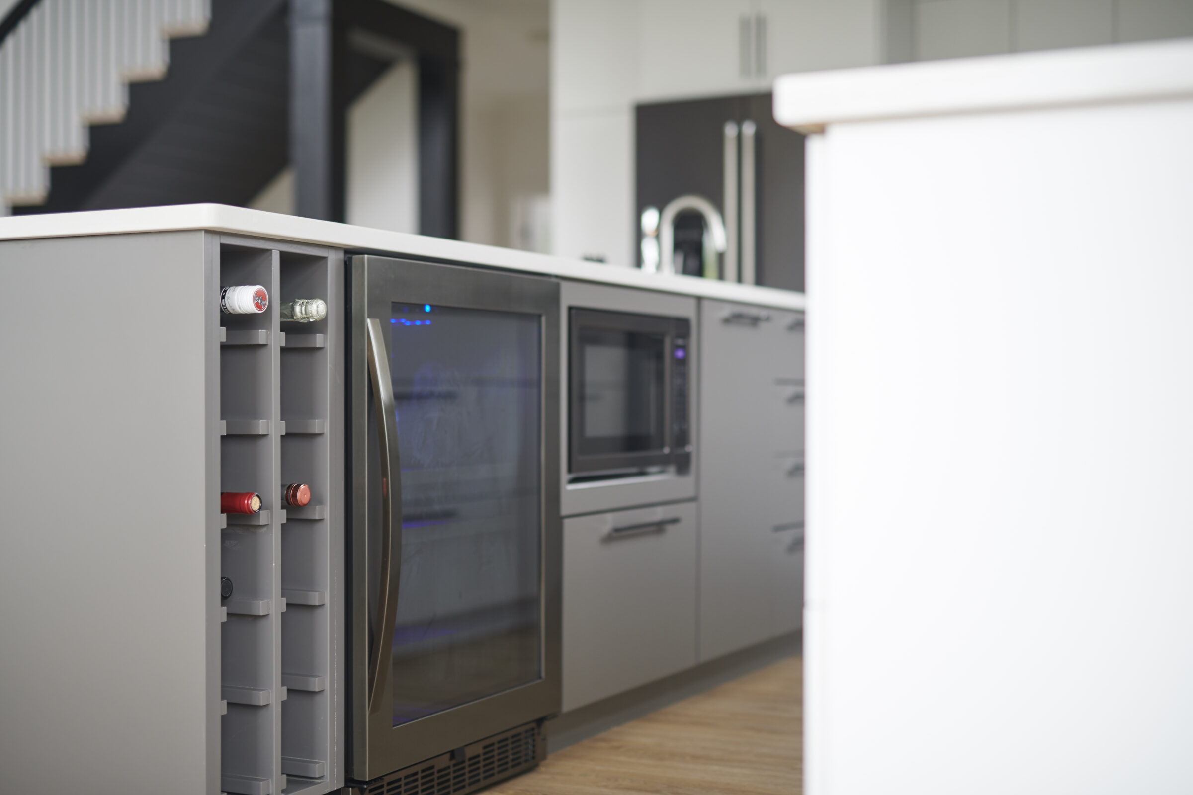 Modern kitchen interior with a wine cooler, built-in ovens, and a staircase. Neutral colors with steel and wood finishes predominate the elegant space.