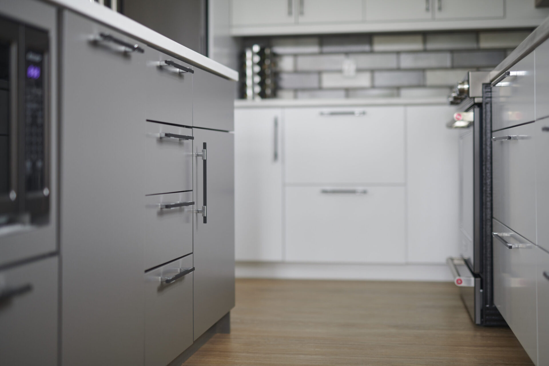 A modern kitchen with gray cabinets, stainless steel handles, white countertops, and a tiled backsplash. Focus is on the foreground with a blurred background.