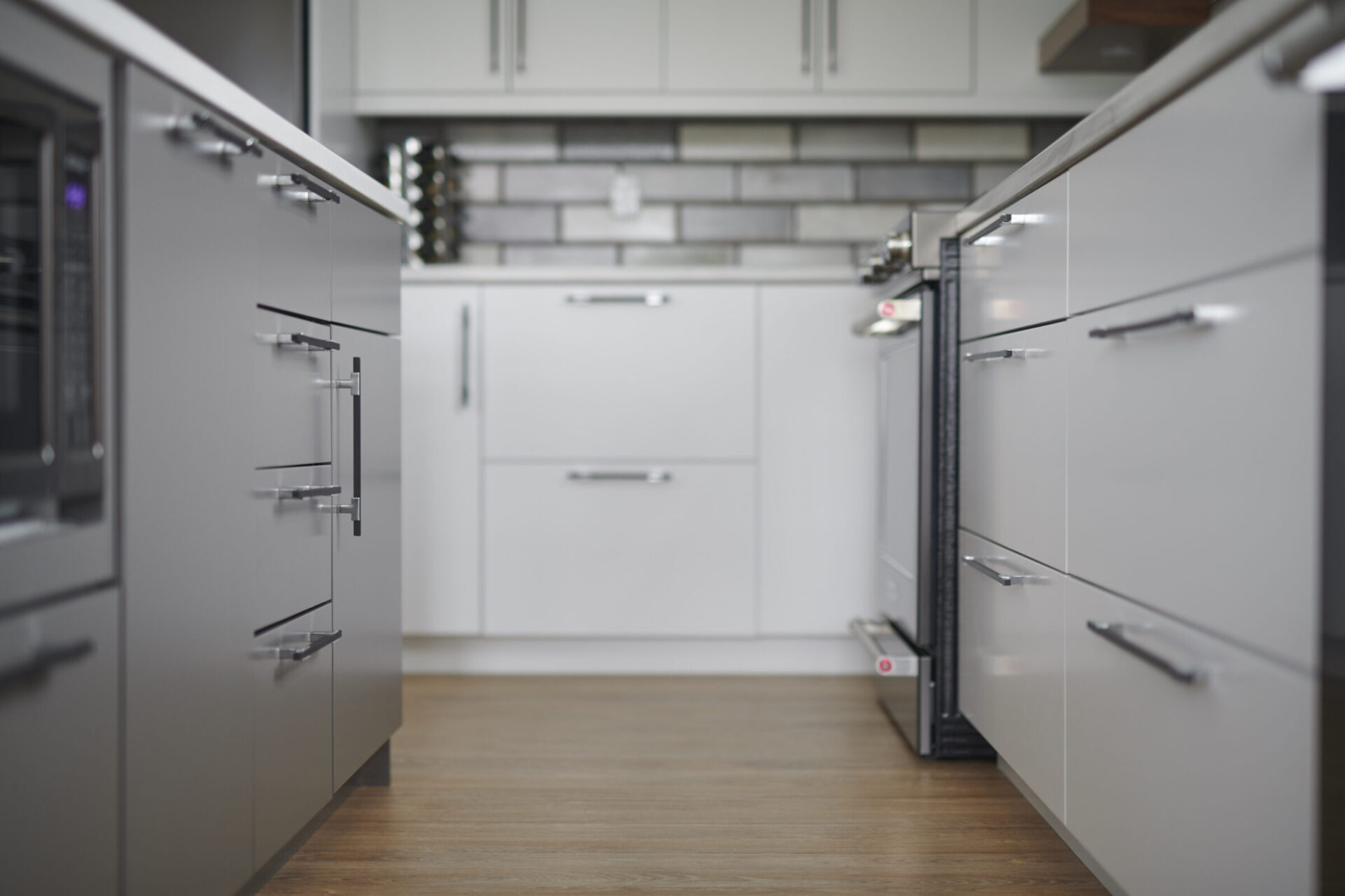 A modern kitchen interior with white cabinetry, gray countertops, stainless steel handles, subway tile backsplash, and wood flooring, taken with a shallow depth of field.