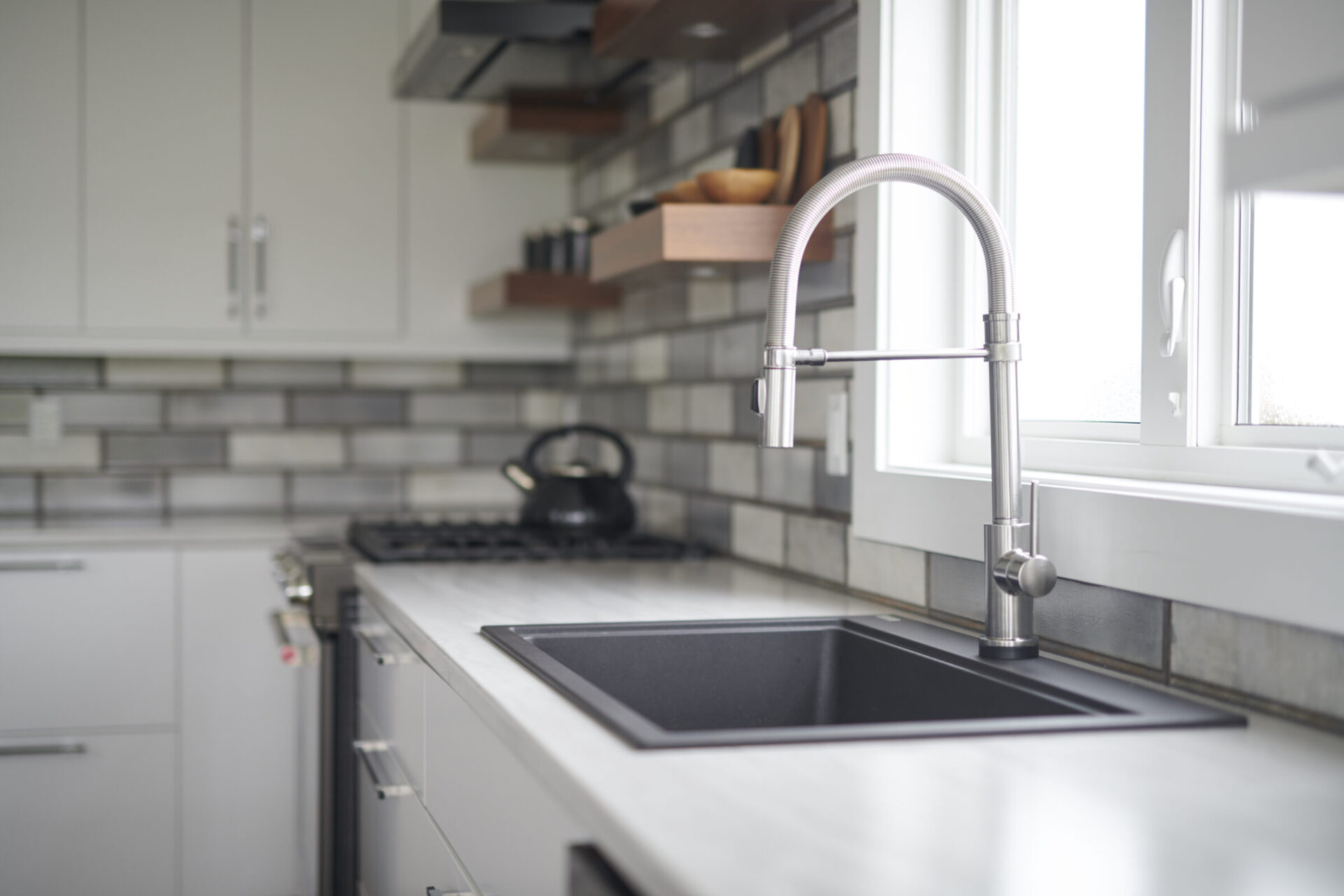 A modern kitchen featuring a stainless steel faucet over a black sink, white cabinets, a tiled backsplash, and a kettle on the stove.
