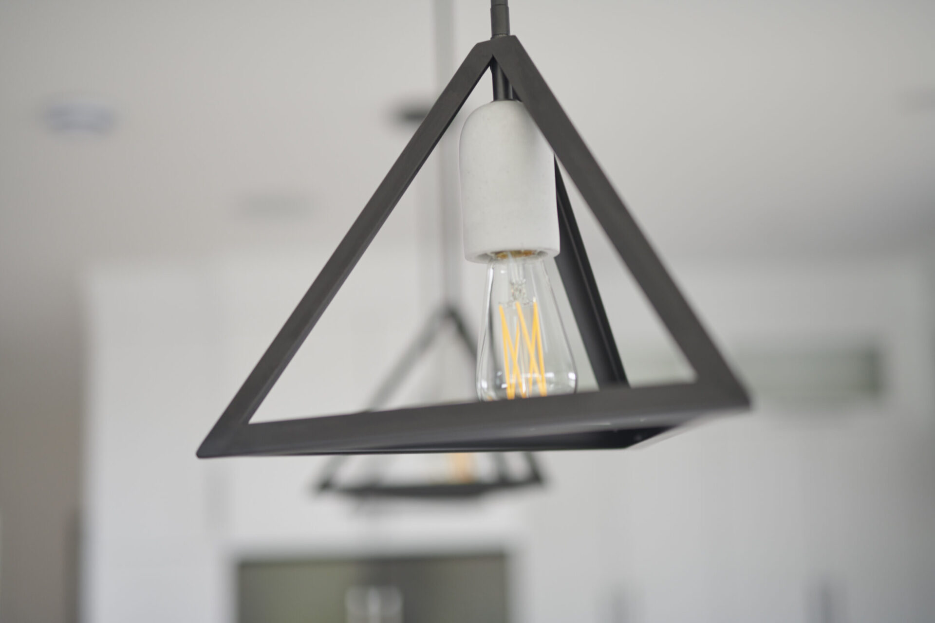 A modern pendant light with a geometric black metal frame hangs indoors. An Edison bulb glows softly against a blurred white interior background.
