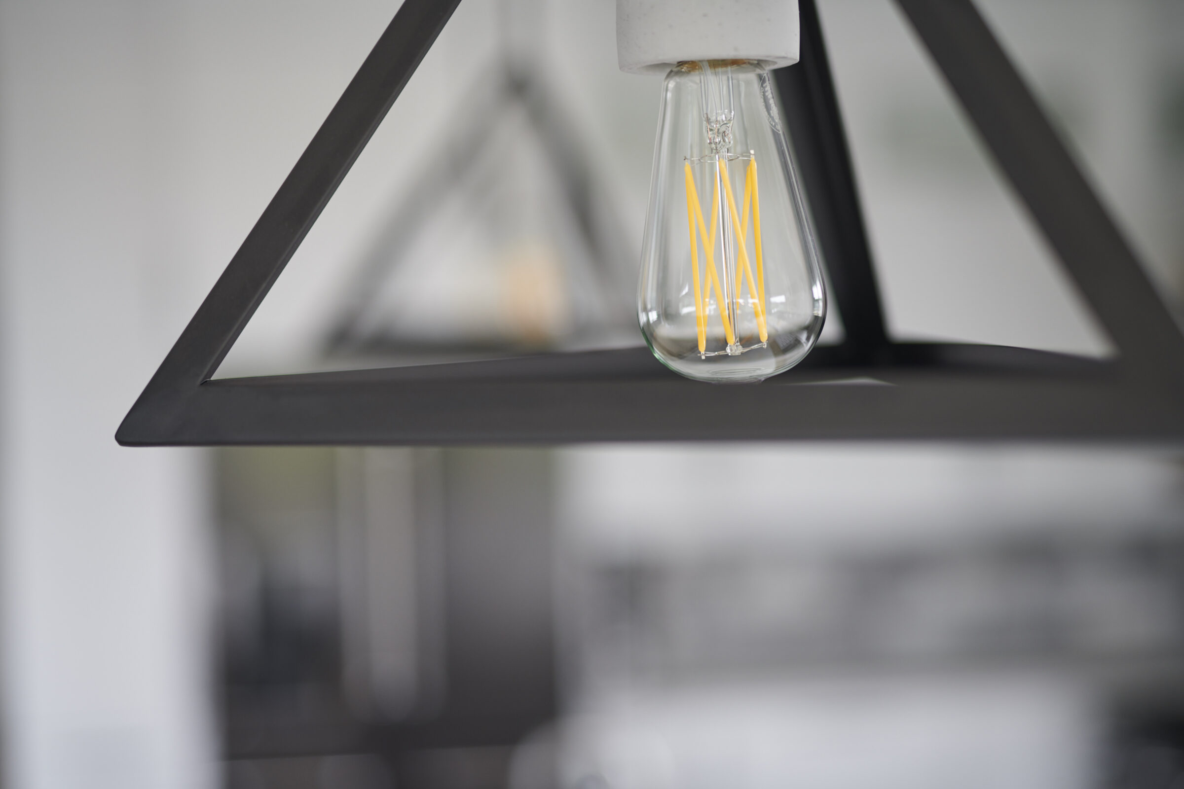Close-up of a modern LED filament light bulb hanging from a geometric black metal lamp frame, with a blurred interior background.