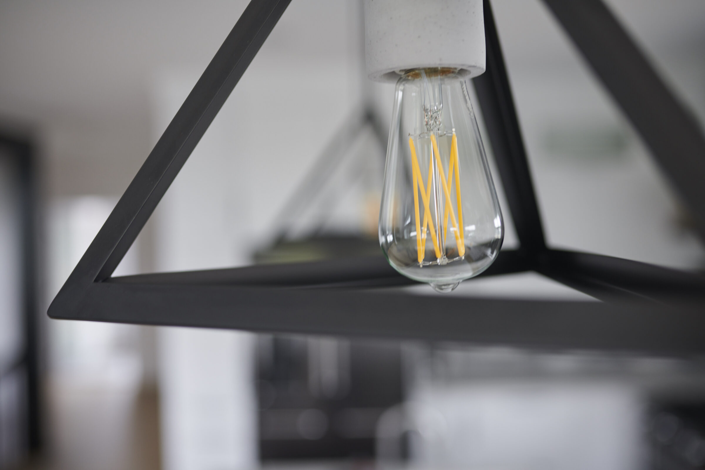 A modern, clear glass light bulb with a visible filament hangs from a gray concrete holder attached to a black geometric frame, blurring the background interior.