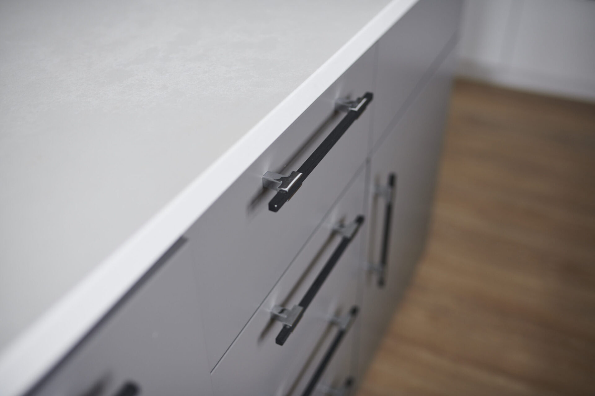 This is a close-up image of a modern kitchen cabinet with a marble countertop, featuring black handles. The focus is on the grey drawers, against a wooden floor background.