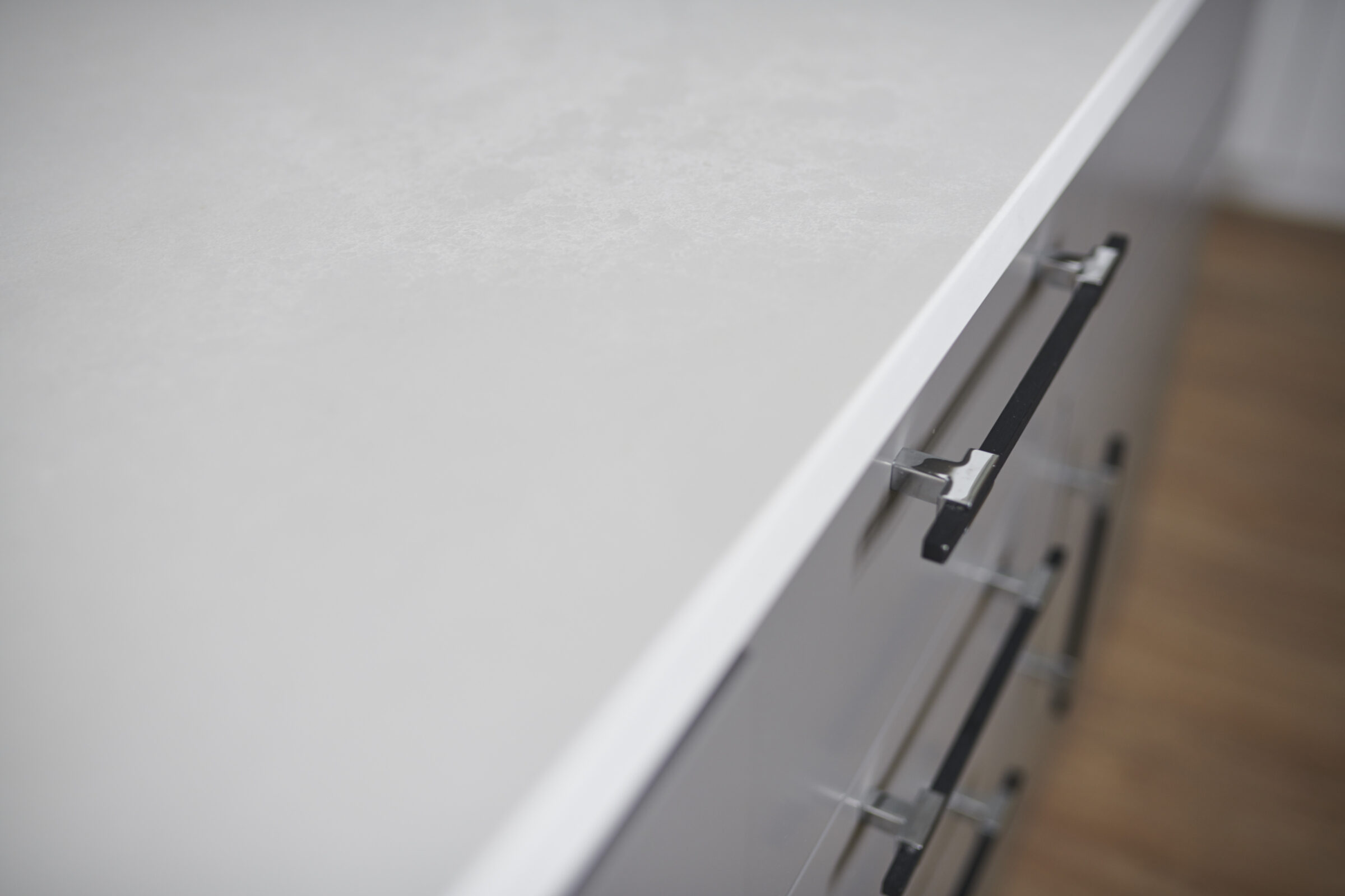The image shows a close-up of a white countertop with a modern, minimalist metal handle on a cabinet or drawer, situated above a wooden floor.