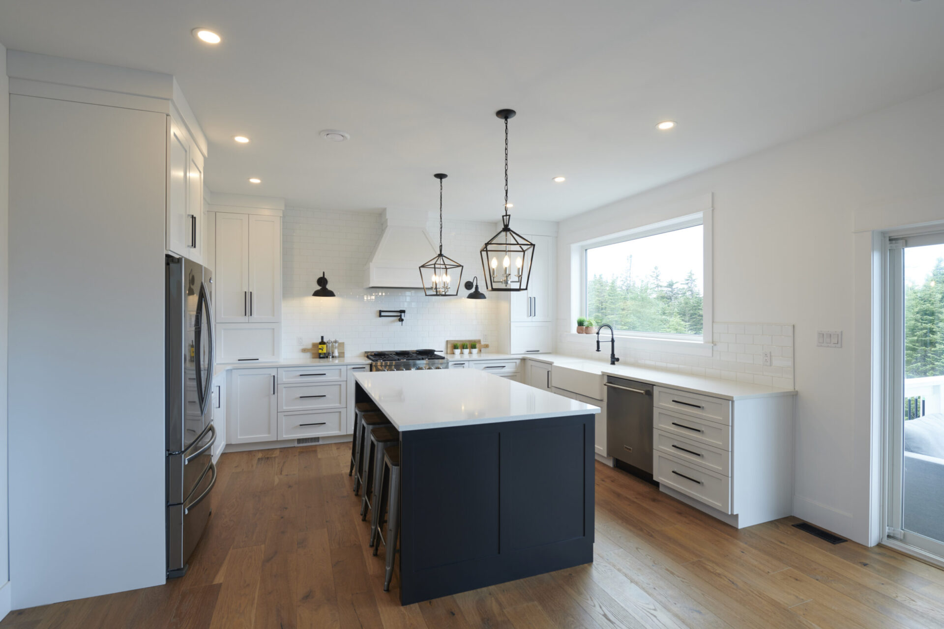 A modern kitchen with white cabinetry, dark island, stainless steel appliances, pendant lights, subway tile backsplash, hardwood floors, and natural light.