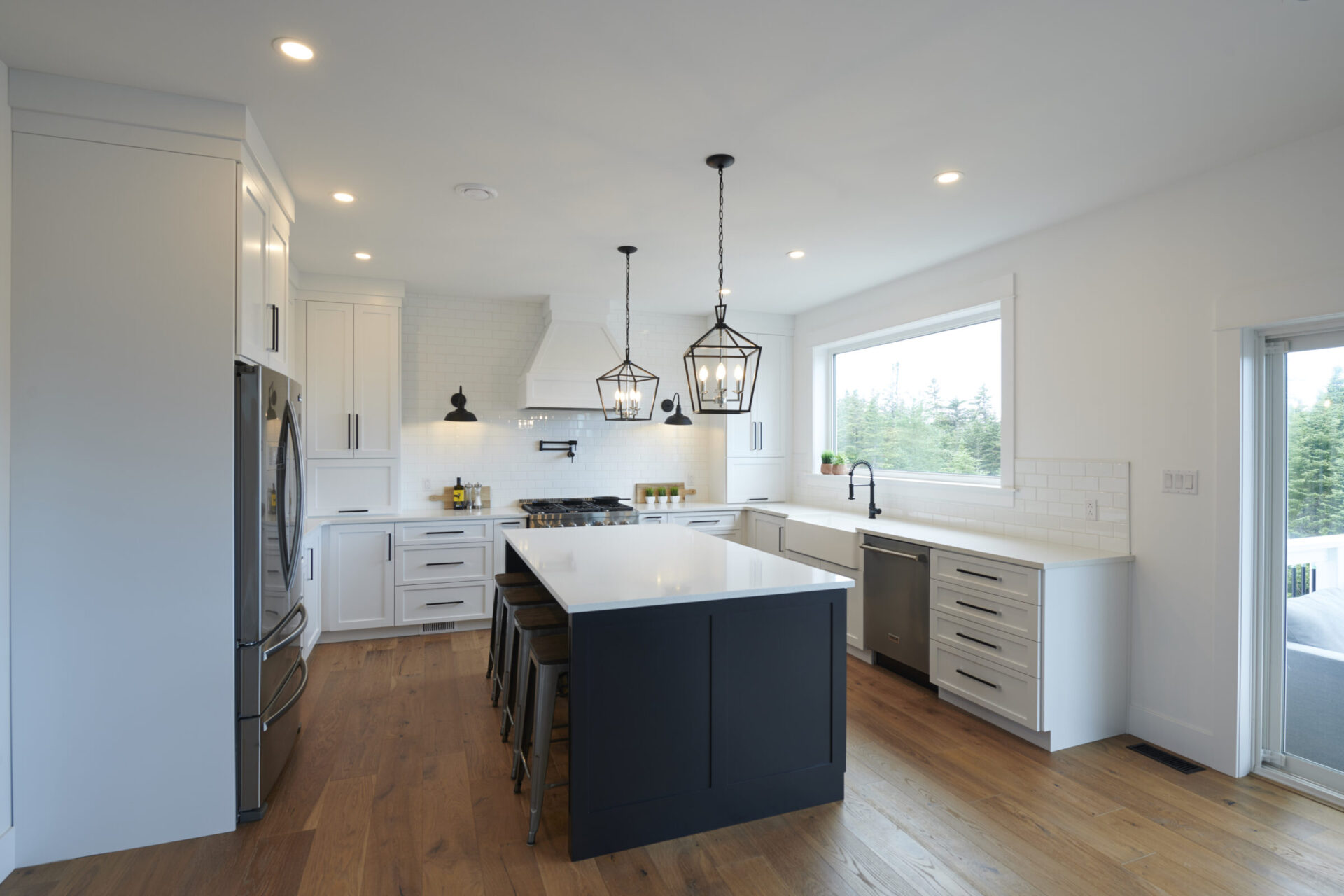 A modern kitchen with white cabinets, stainless steel appliances, dark blue island, pendant lights, subway tiles, hardwood floors, and a bright window.