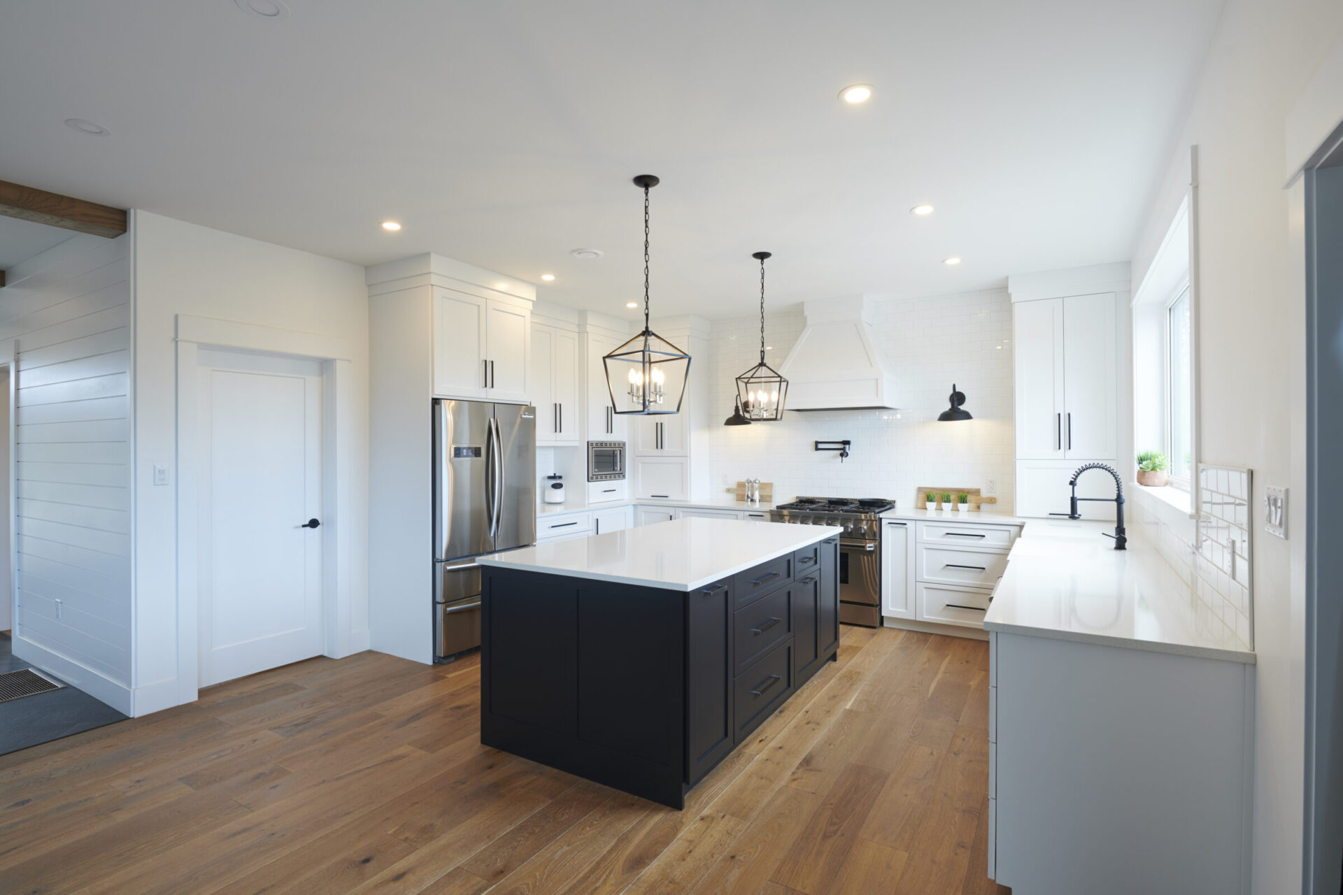A spacious modern kitchen with white cabinets, a central black island, stainless steel appliances, hardwood floors, and pendant lighting fixtures.