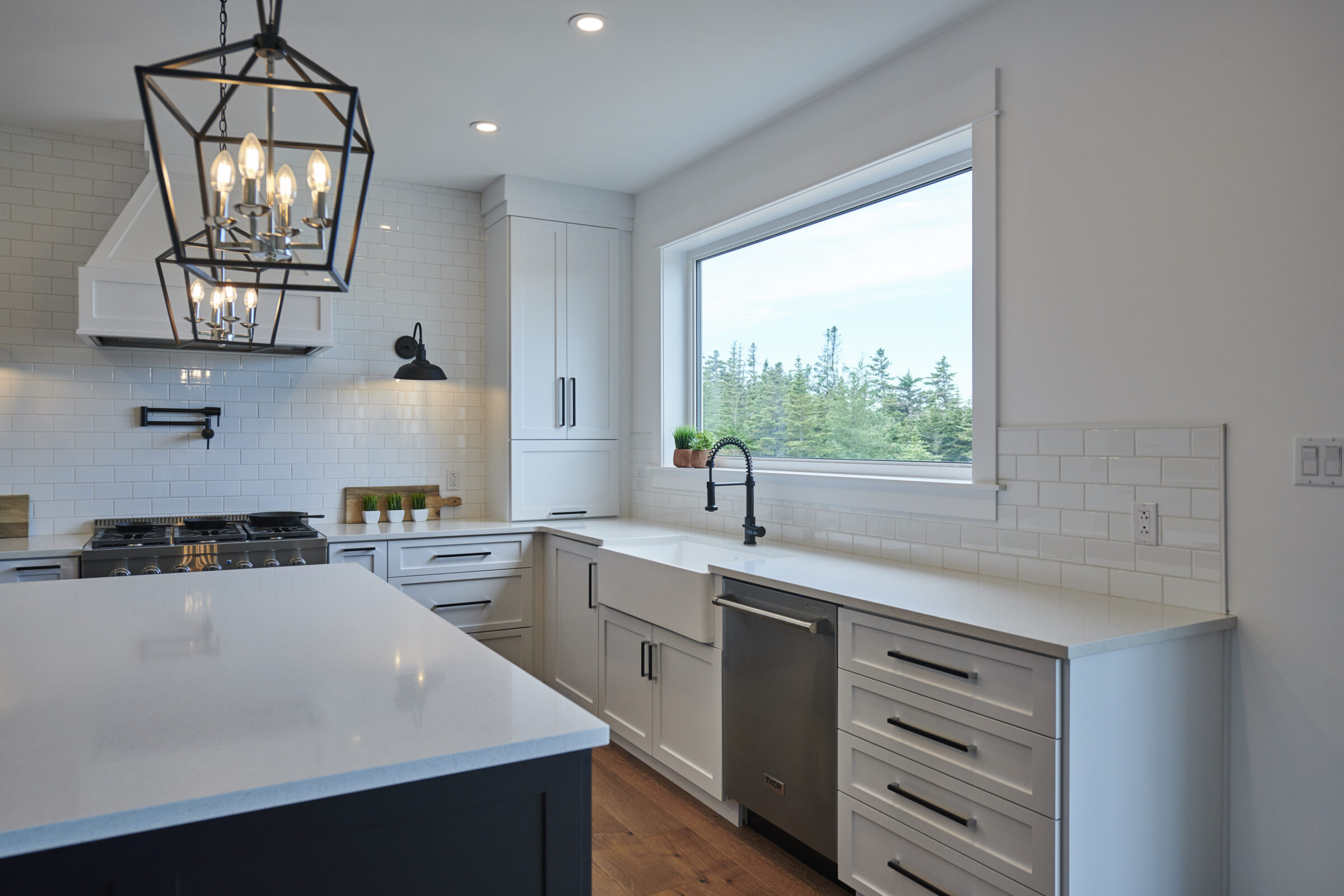Modern kitchen with white cabinets, subway tile backsplash, large window with forest view, geometric pendant light, and stainless steel appliances.