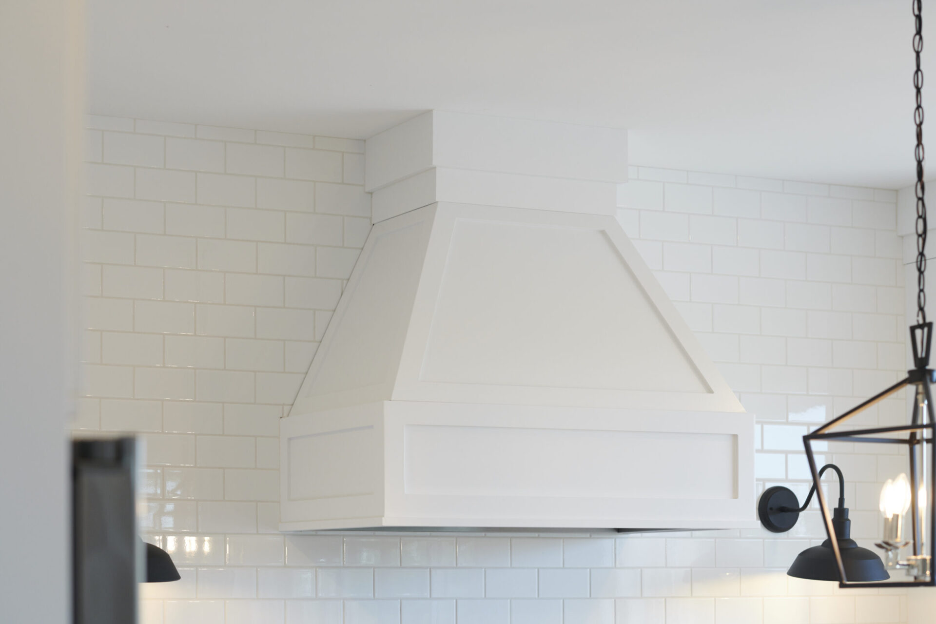 A modern kitchen with white subway tiles features a large white range hood. A black pendant light with an exposed bulb hangs to the right.